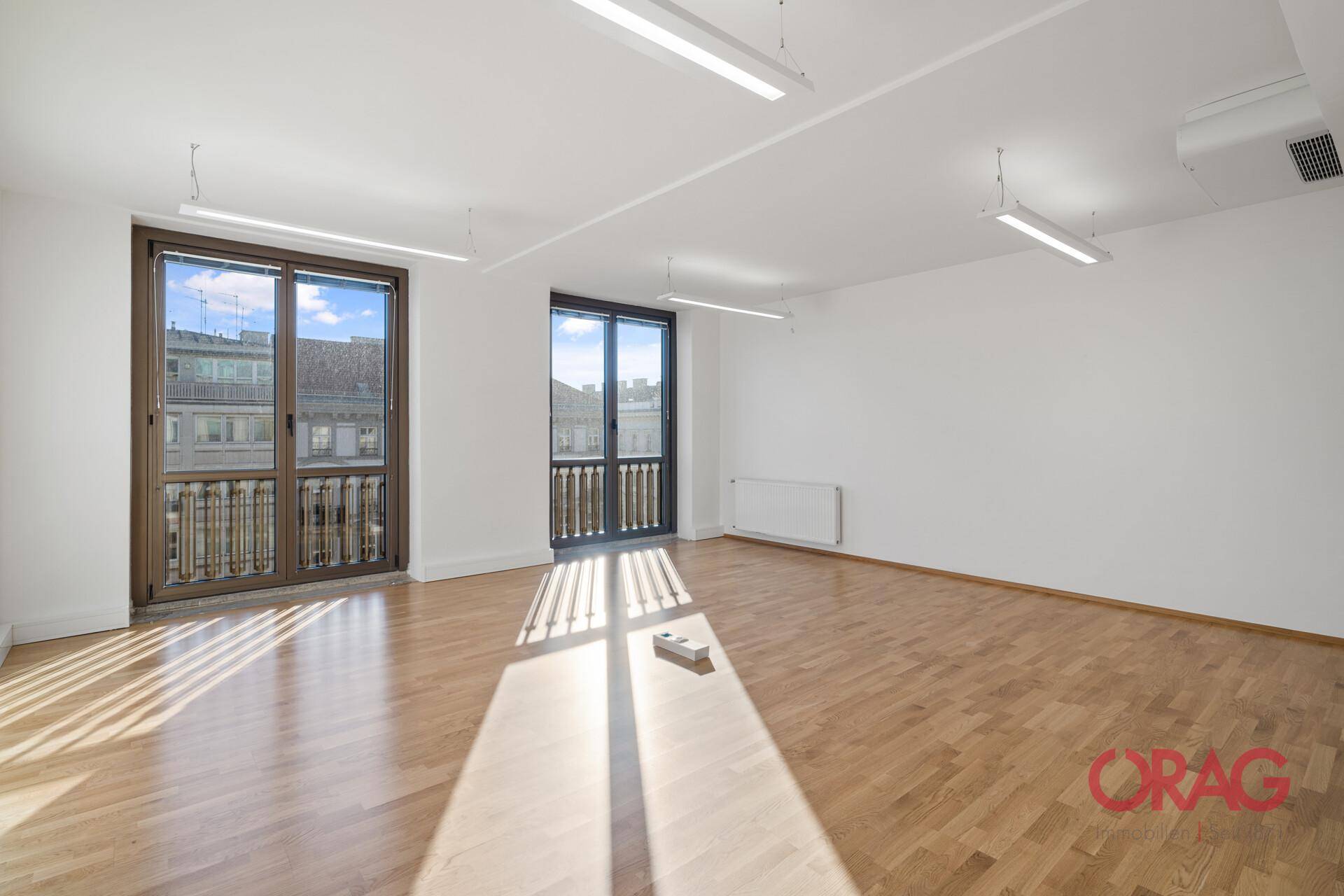 Geräumiger Büroraum mit Holzboden und zwei großen Fenstertüren, die Zugang zu kleinen französischen Balkonen bieten.