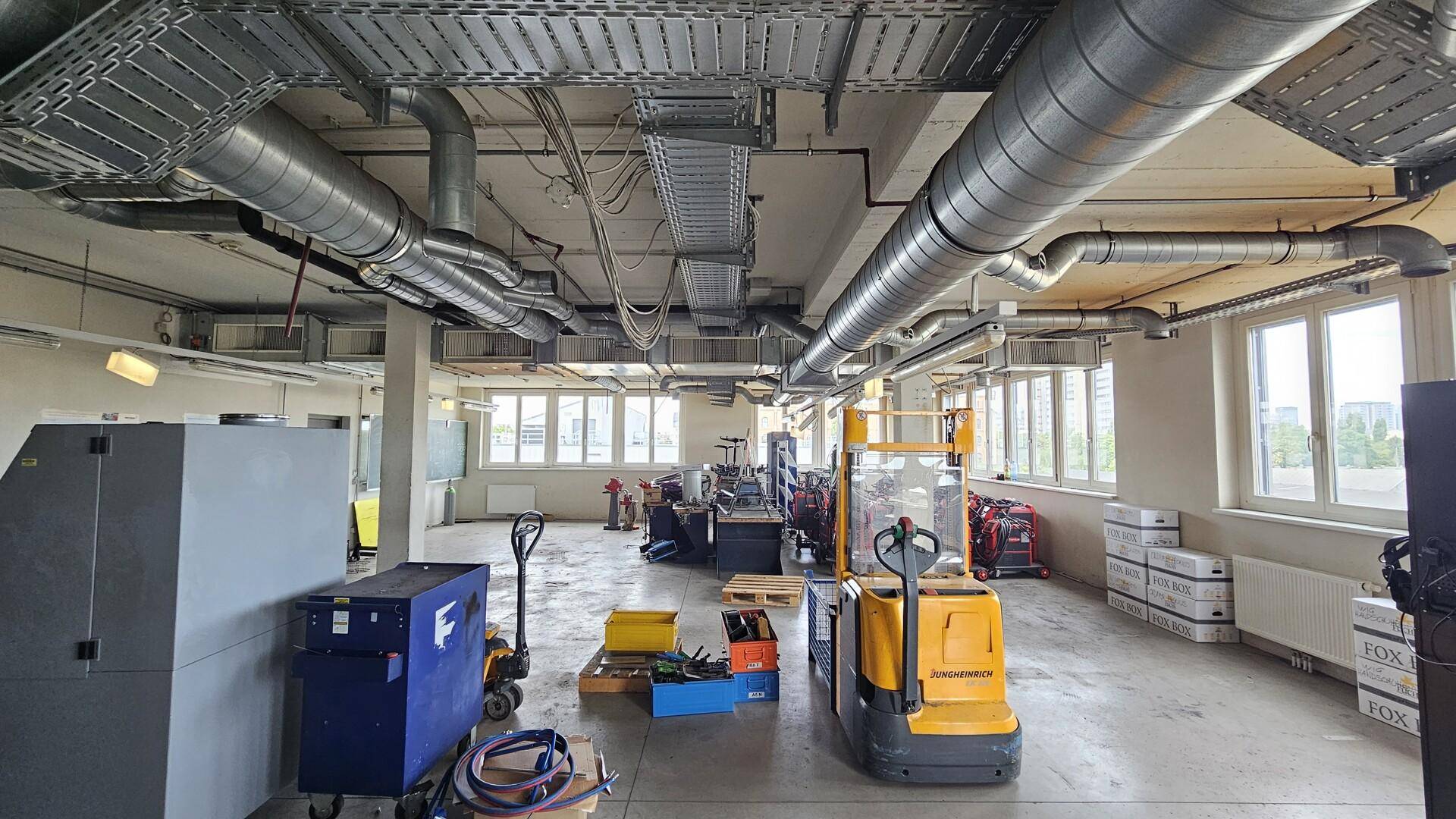 Großer Industrieraum mit sichtbaren Lüftungsrohren, Maschinen und vielen Fenstern.