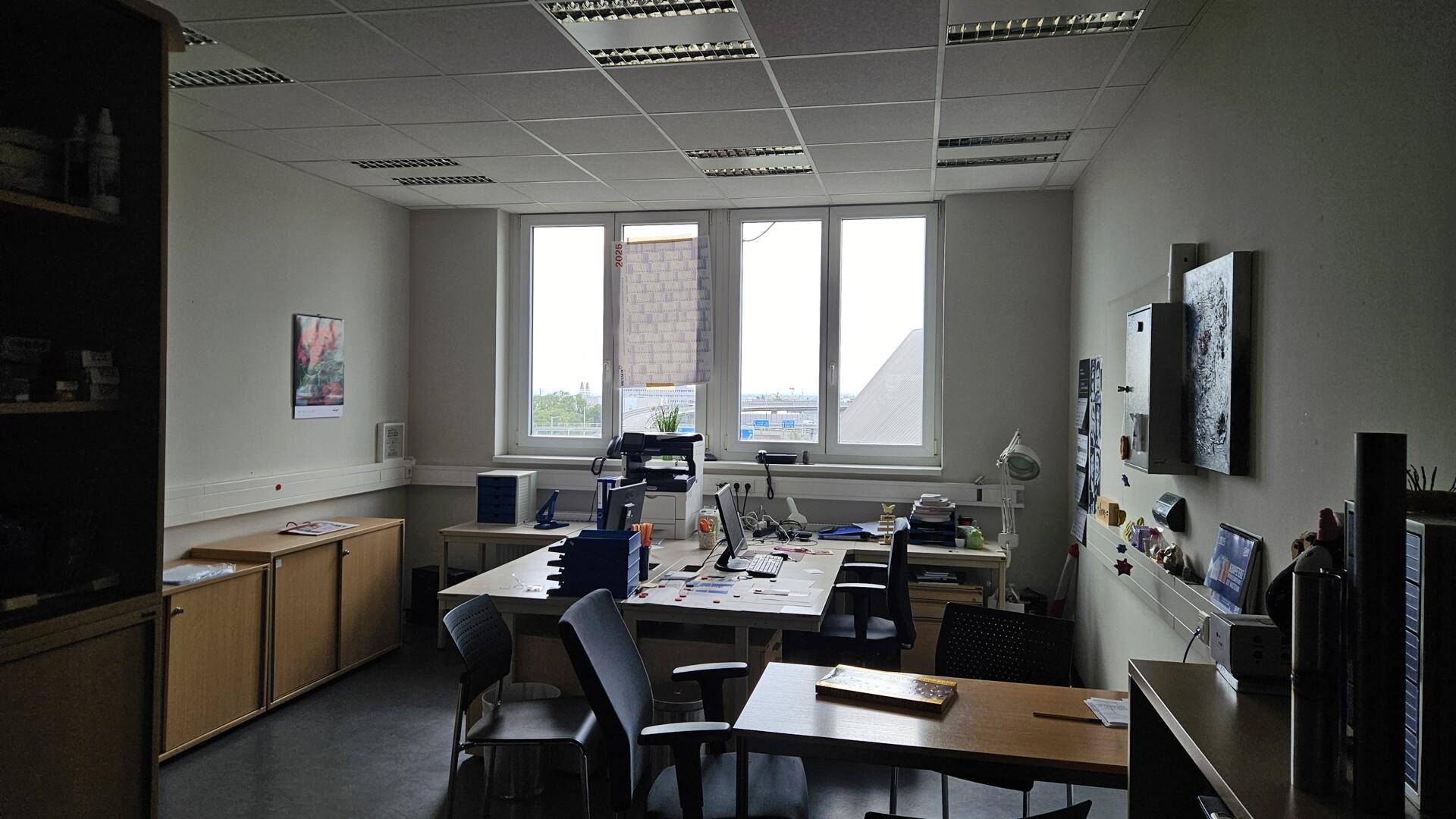 Möbliertes Büro mit Schreibtischen, Stühlen und Schränken, hell durch große Fenster.