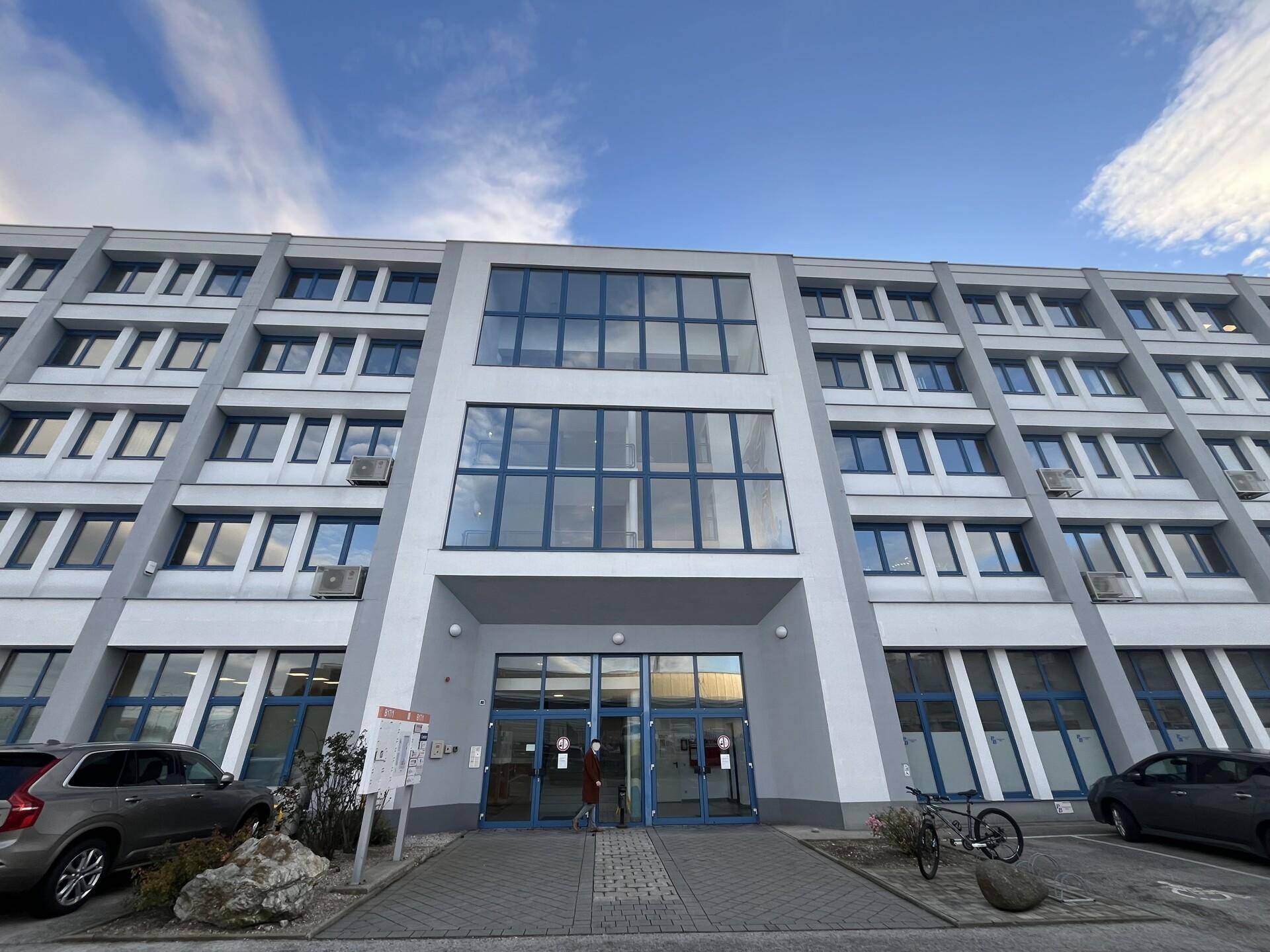 Frontansicht eines modernen Bürogebäudes mit großem Eingangsbereich und vielen Fenstern unter blauem Himmel.