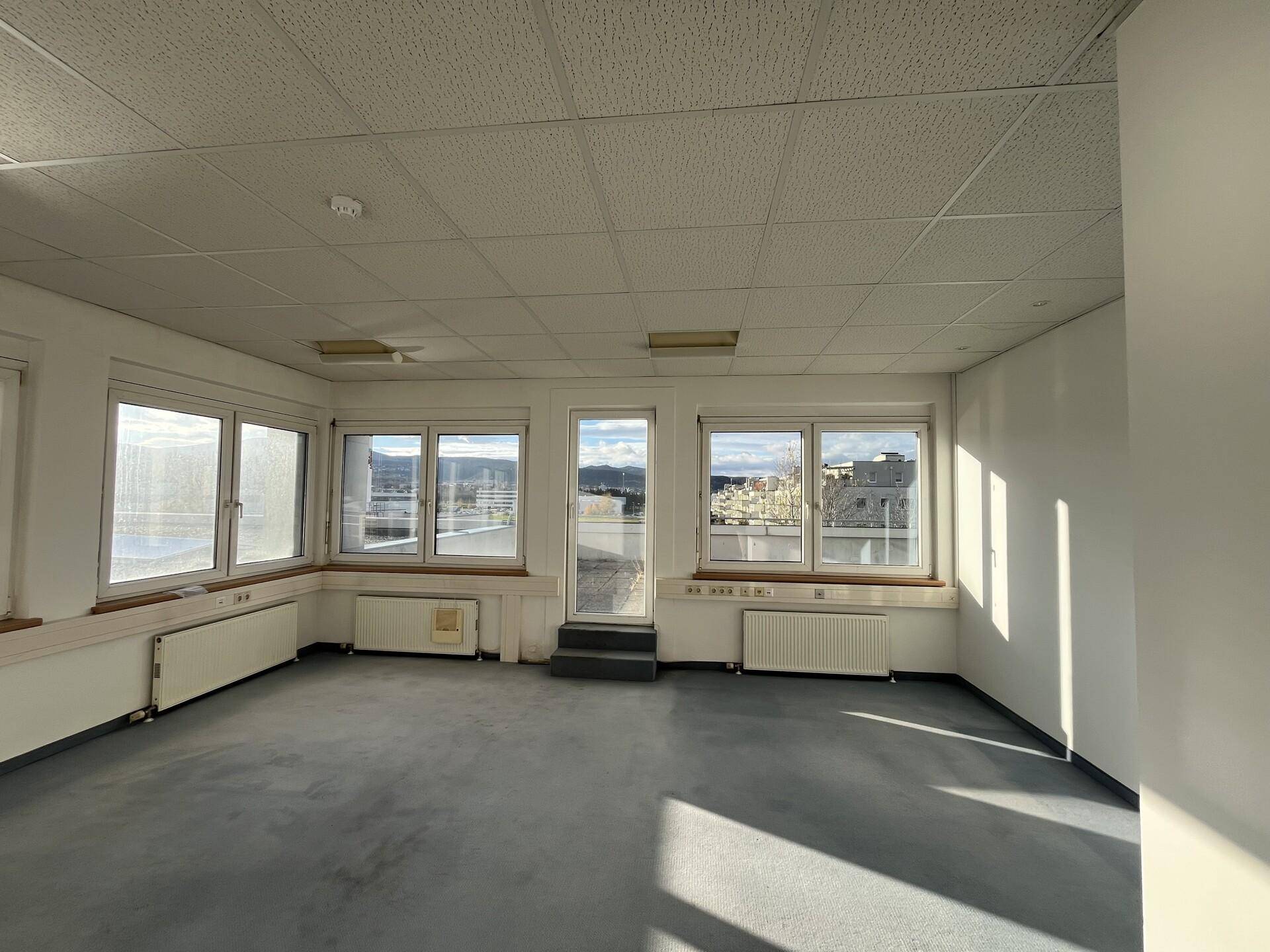 Heller Büroraum mit vielen Fenstern und einer Tür, die auf eine Terrasse oder einen Balkon führt.
