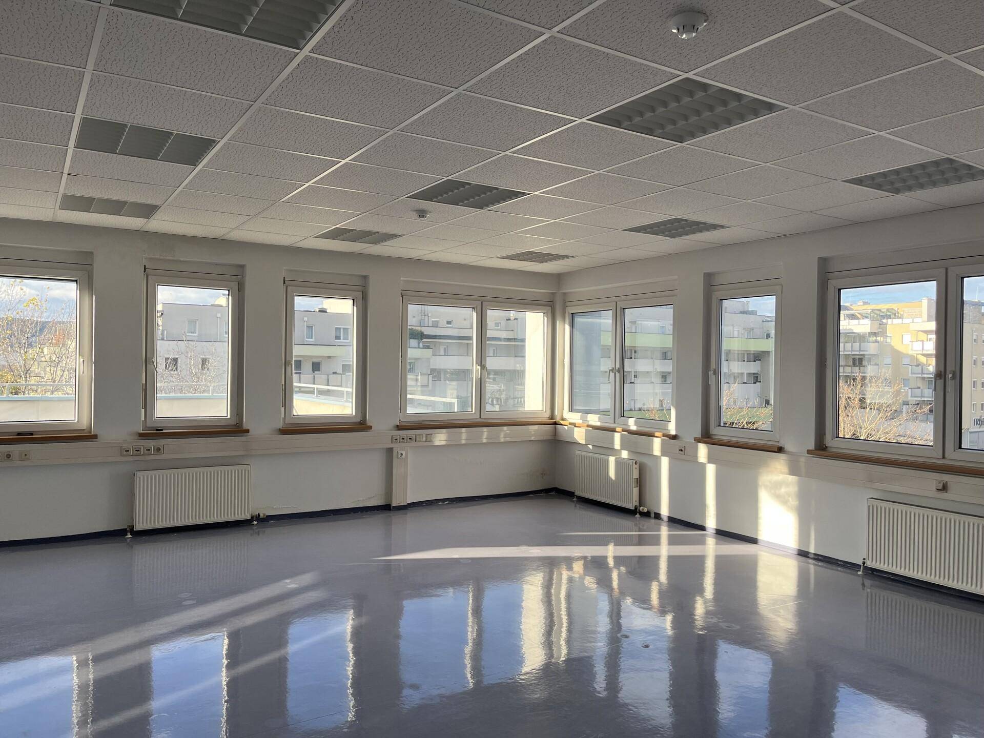 Geräumiges, leeres Büro mit vielen Fenstern, die viel Tageslicht hereinlassen, und einem hellen Boden.
