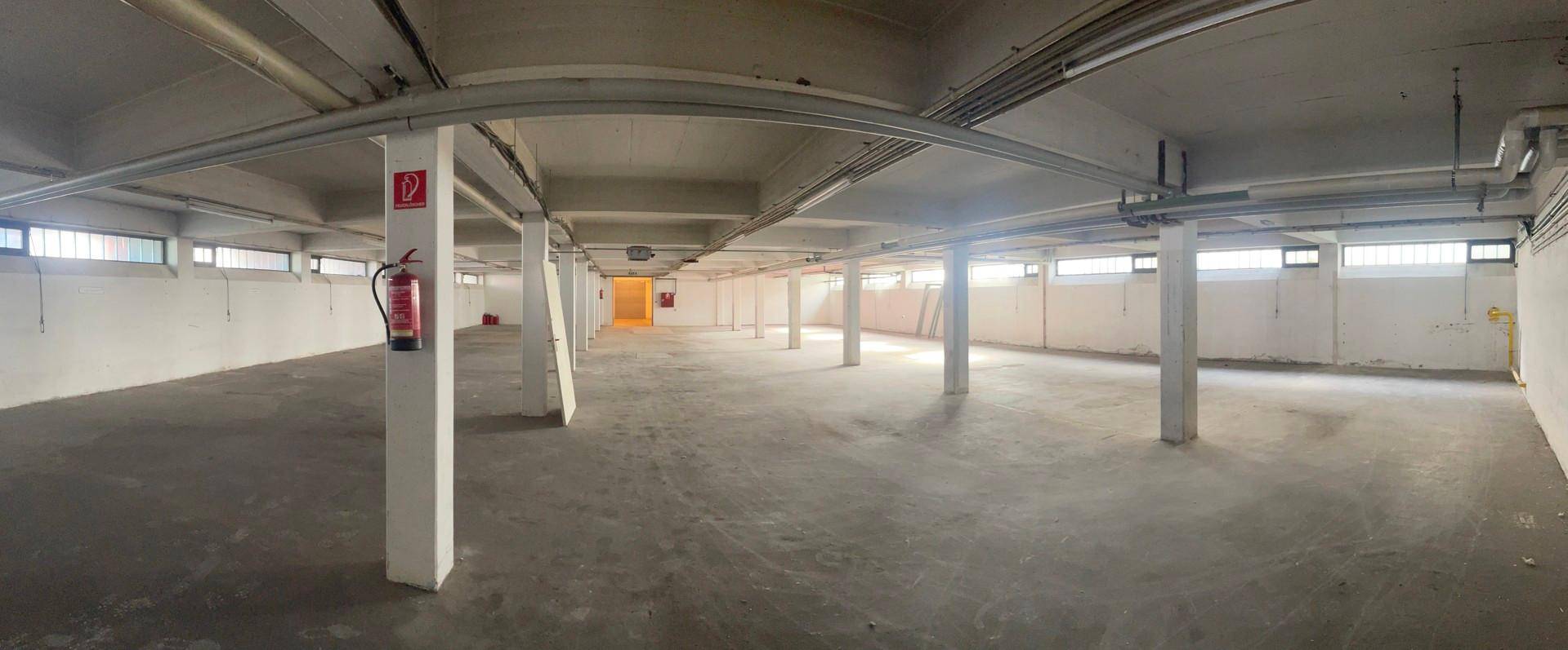 Panoramaansicht eines weitläufigen Lagerraums mit Betonboden, Säulen und Fenstern entlang der Wände.