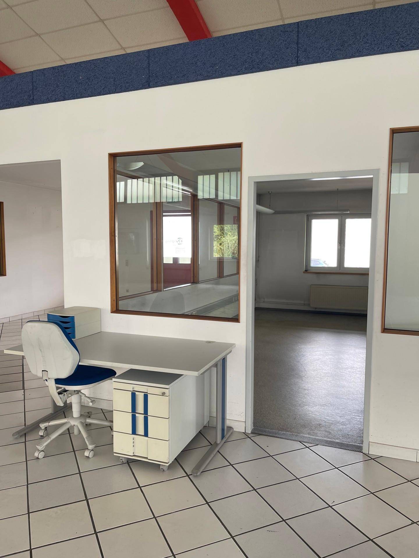 Bürobereich mit Schreibtisch und Stuhl, Blick in einen weiteren Raum, beide mit Fliesenboden.