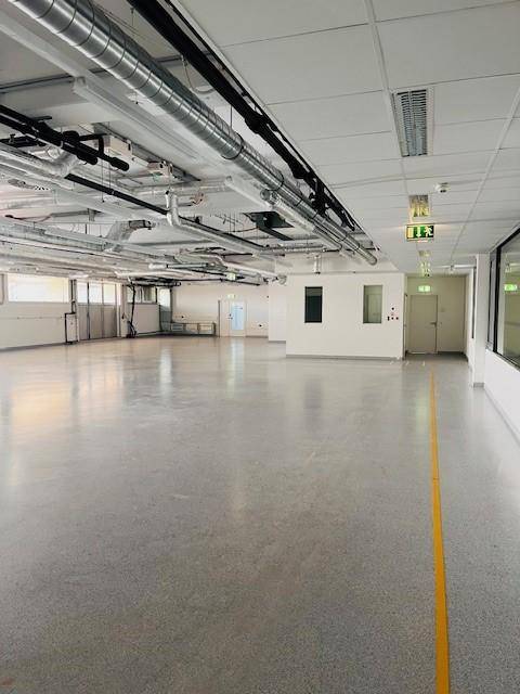 Große, leere Halle mit Industrieboden, sichtbaren Lüftungsrohren und hohen Decken.