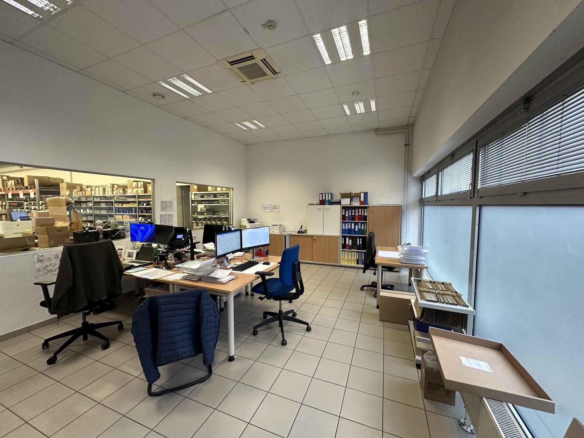Modernes Büro mit mehreren Arbeitsplätzen, großen Fenstern und Blick auf einen angrenzenden Lagerbereich.