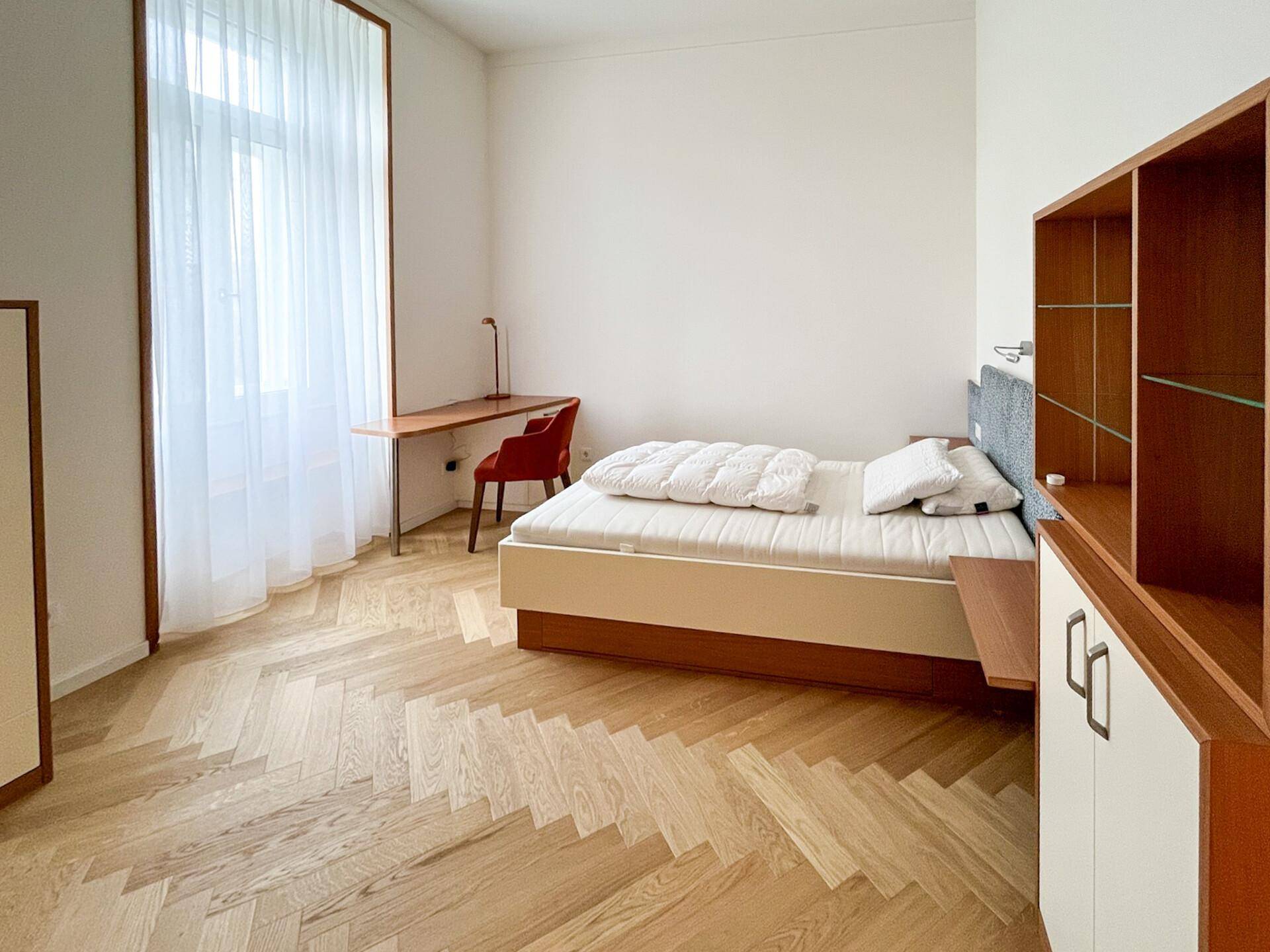 Schlafzimmer mit Fischgrätparkett, großem Fenster und modernem Bett mit Nachttisch.