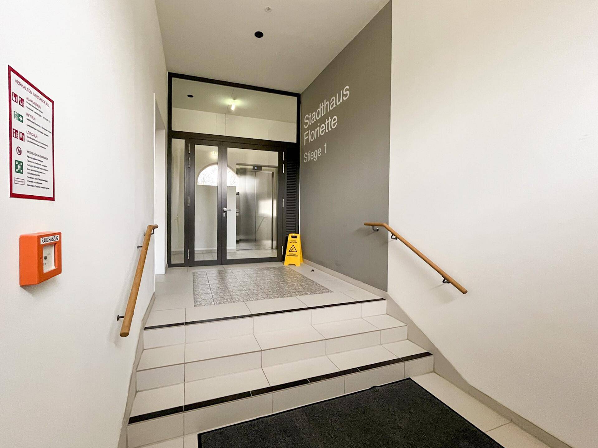 Heller Eingangsbereich des Gebäudes mit Treppe, Handläufen und Zugang zum Aufzug.