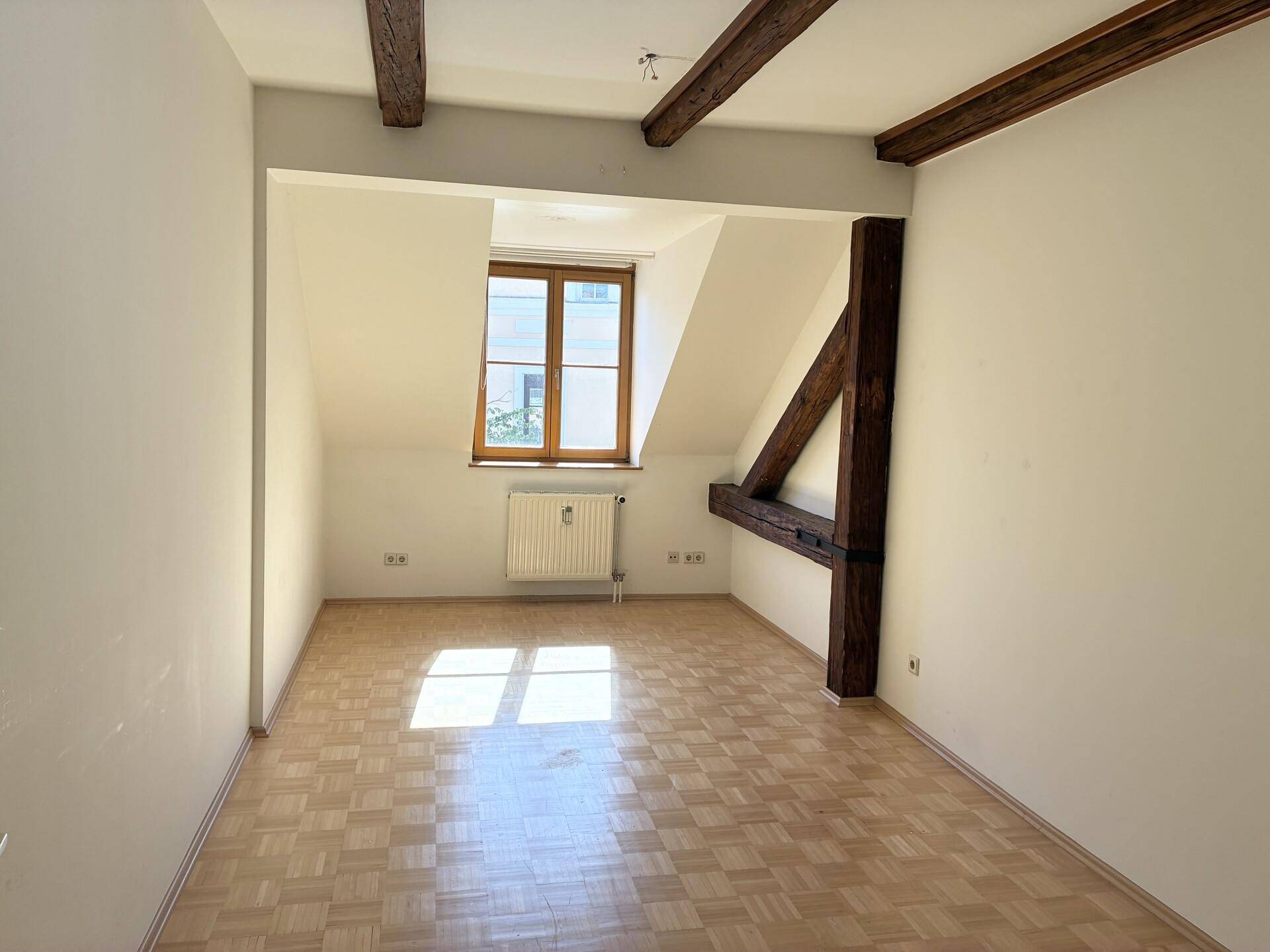 Lichtdurchfluteter Raum im Dachgeschoss mit sichtbaren Holzbalken und Parkettboden.