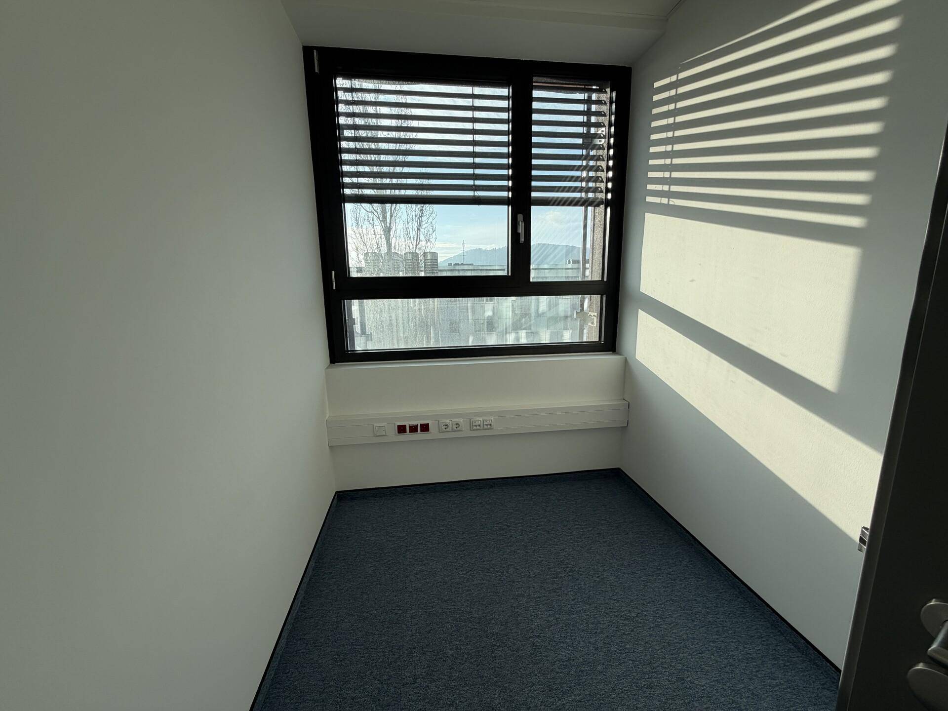 Einzelbüro mit Teppichboden und einem Fenster mit Jalousien, das Tageslicht hereinlässt.