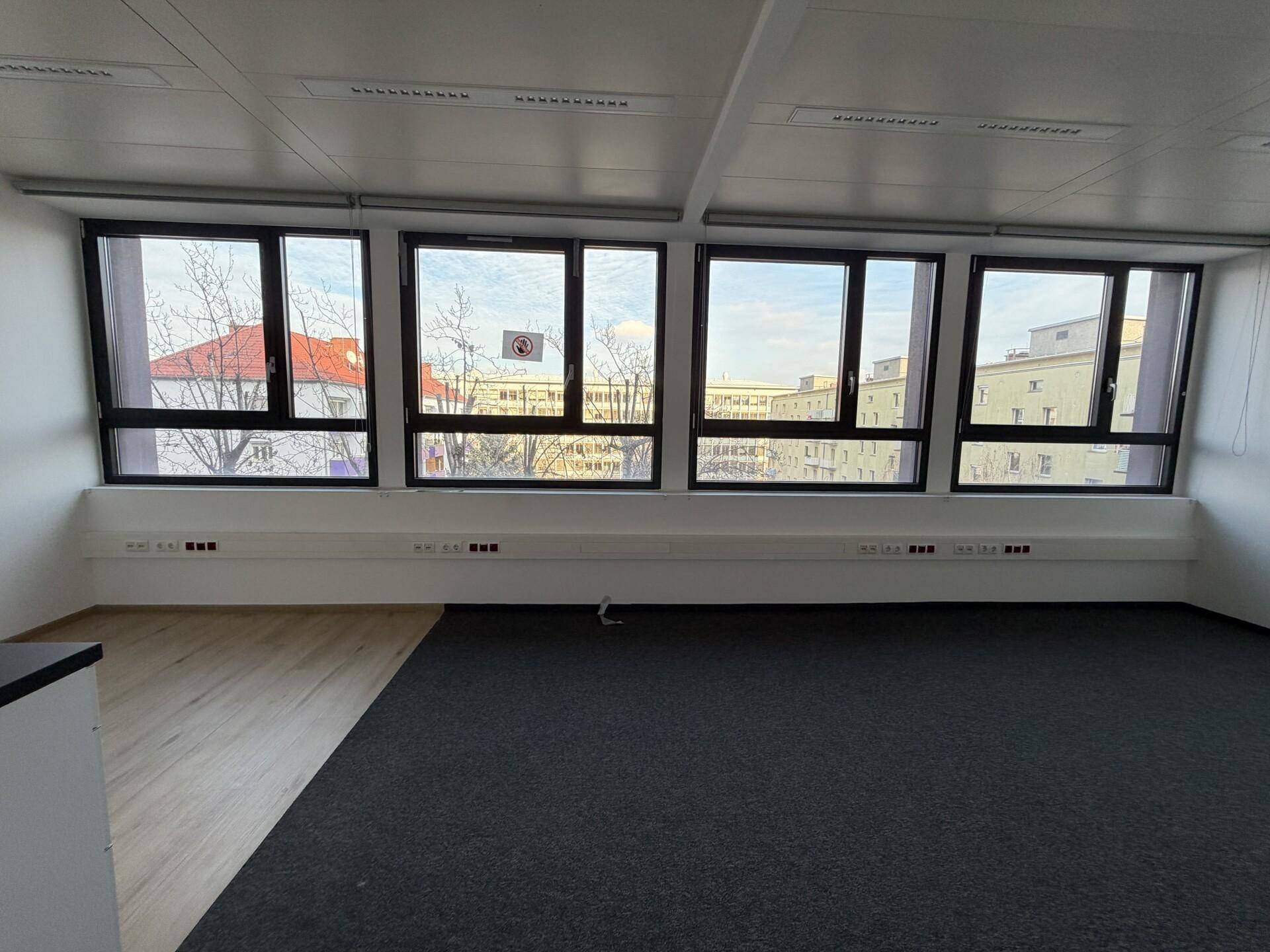 Büroraum mit Teppich- und Laminatboden, großen Fenstern und Blick auf die Stadt.