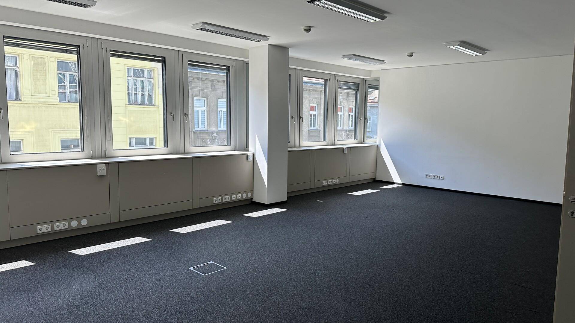 Großes, leeres Büro mit Teppichboden, vielen Fenstern und weiß gestrichenen Wänden.