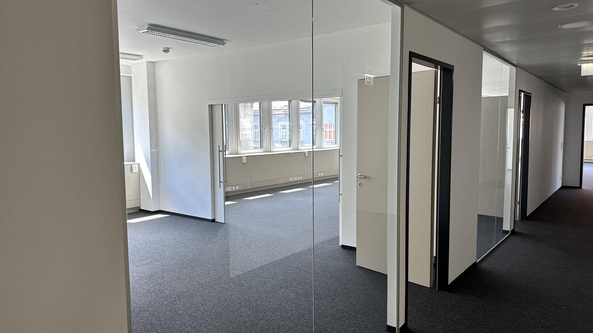 Blick in ein helles Büro durch eine Glastrennwand, mit dunklem Teppichboden und großen Fenstern.