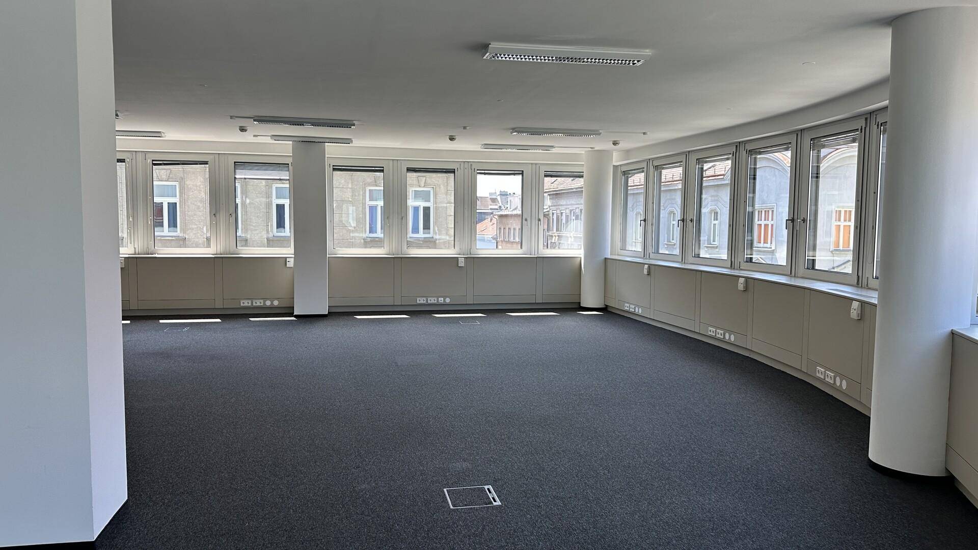 Großes, helles Büro mit gebogener Fensterfront, dunklem Teppichboden und viel natürlichem Lichteinfall.