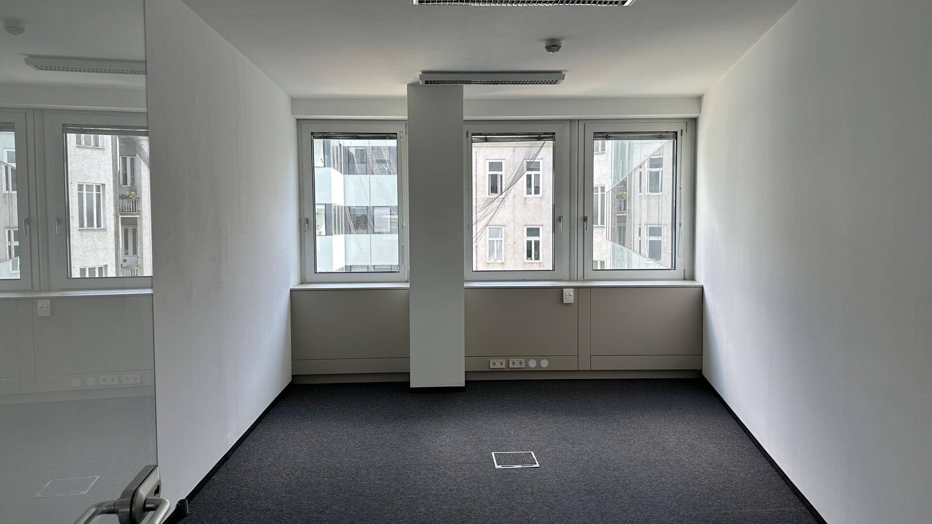Kleineres, helles Büro mit dunklem Teppichboden und drei Fenstern, die den Raum mit Licht füllen.