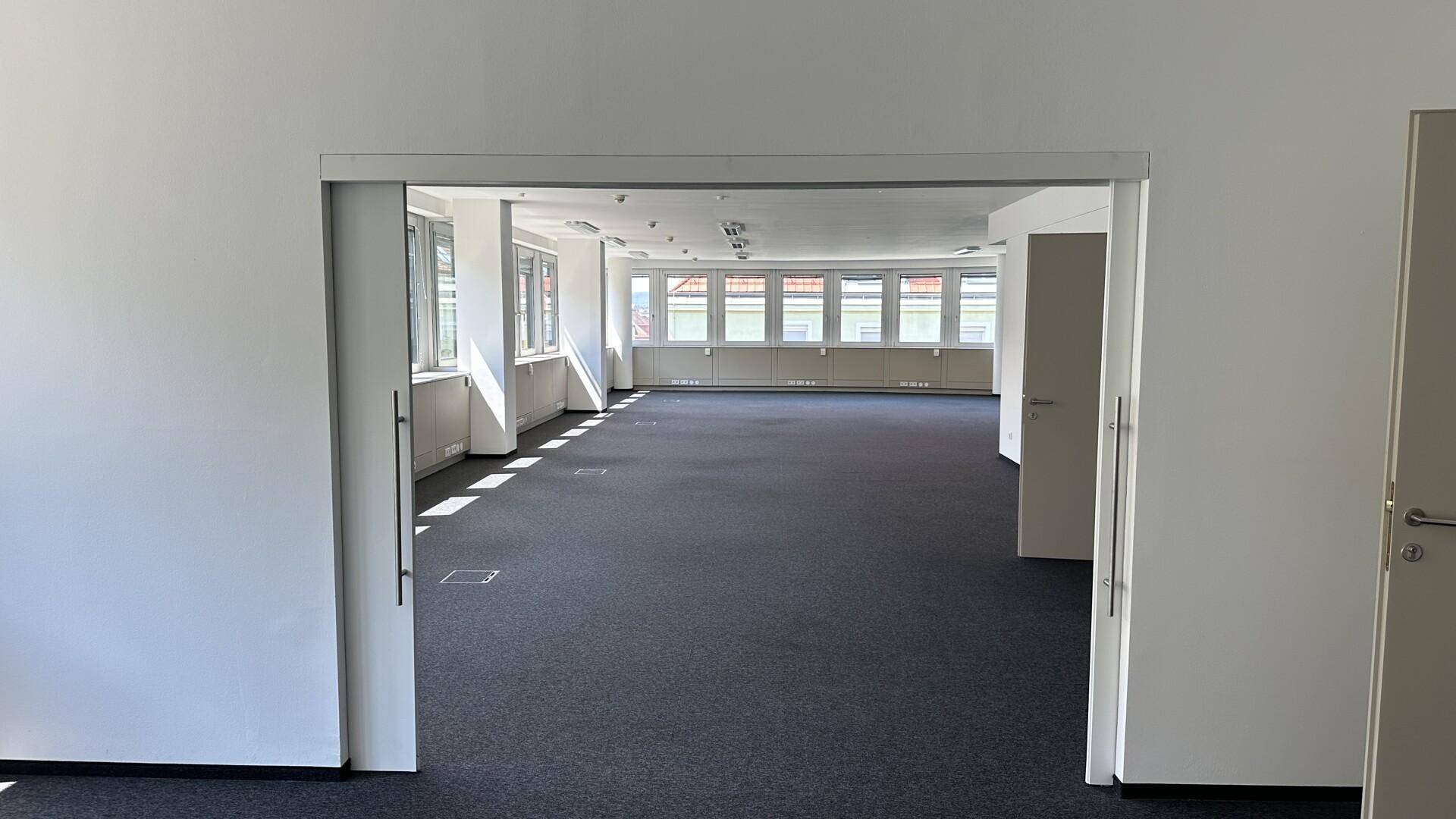 Weitläufiger, leerer Büroraum mit dunklem Teppichboden und zahlreichen Fenstern, die den Raum erhellen.