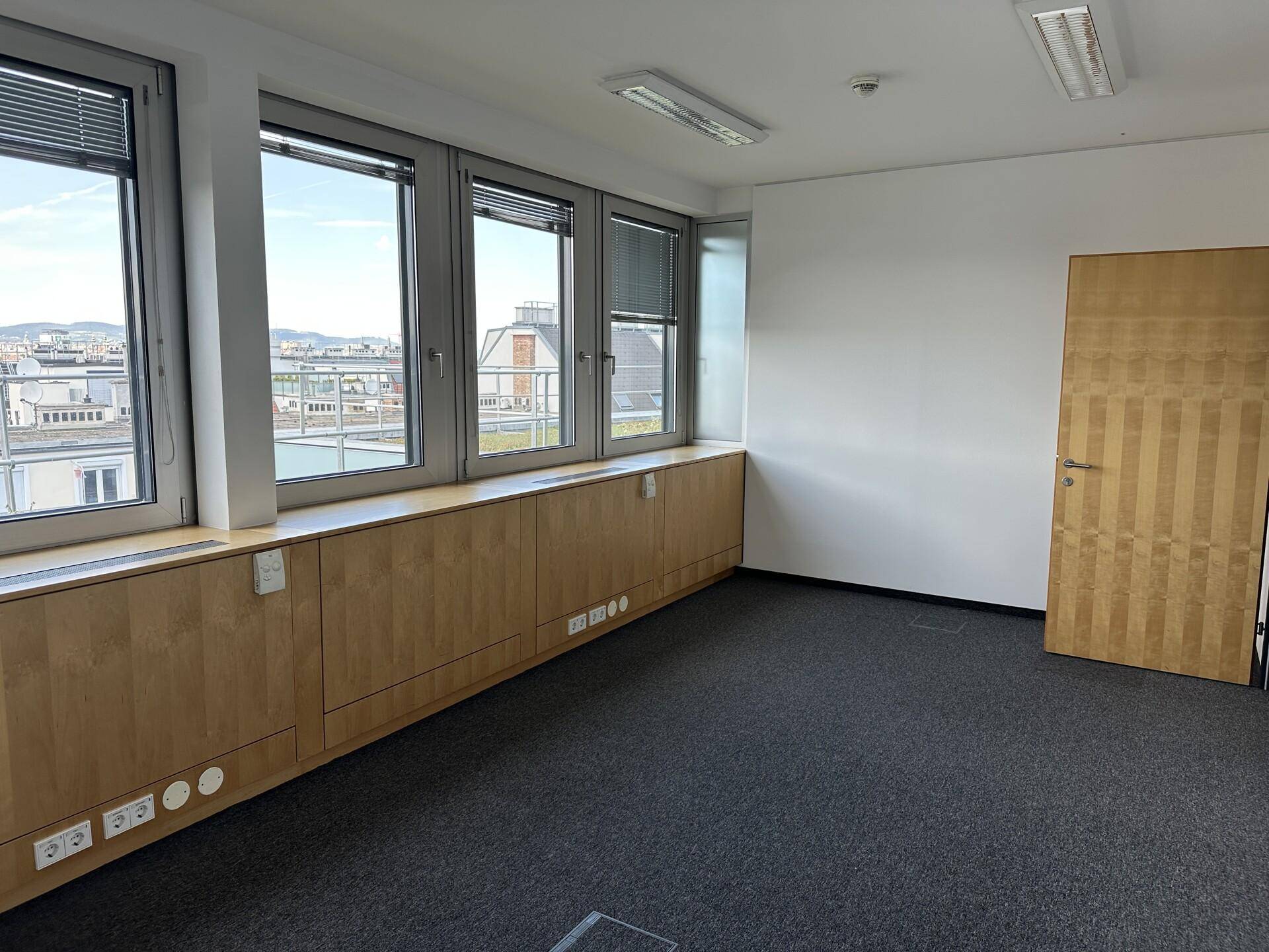 Leeres Büro mit Teppichboden, Holzverkleidung und weitem Blick über Dächer und Landschaft.