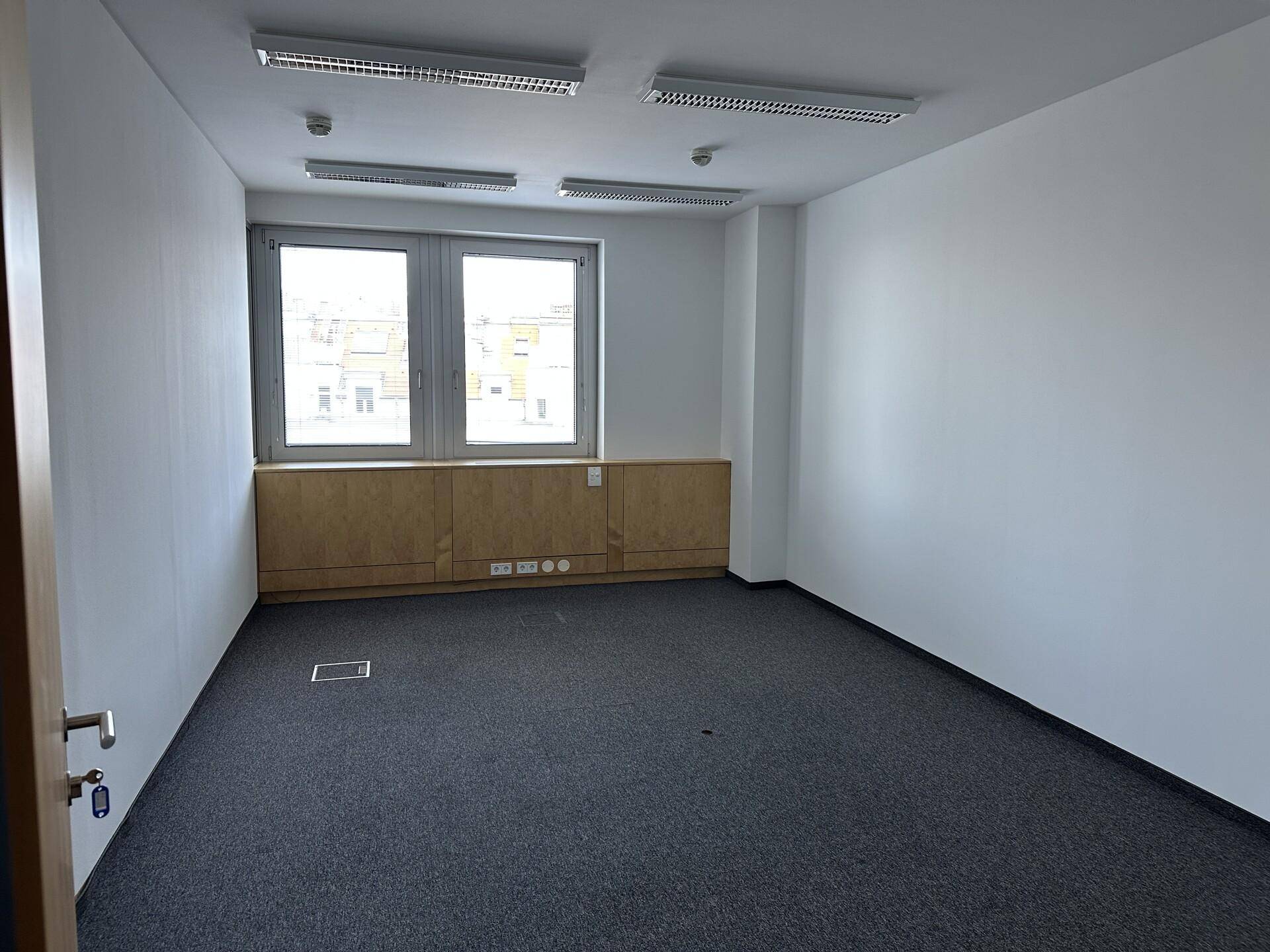 Kleineres, leeres Büro mit zwei Fenstern, Teppichboden und integrierten Holzschränken.
