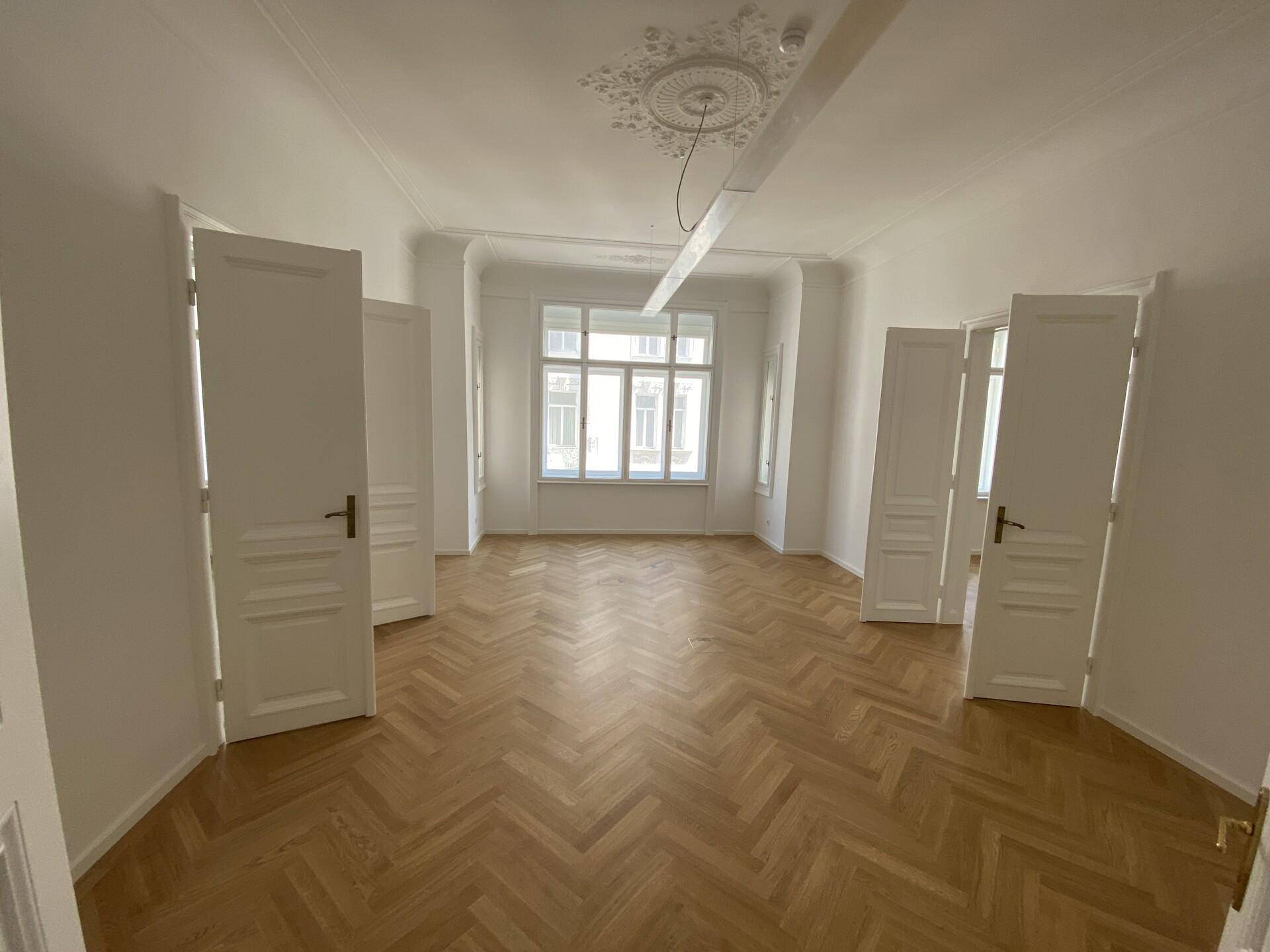 Geräumiges Zimmer mit Fischgrätparkett, Erkerfenster und mehreren weißen Türen, das vielseitig nutzbar ist.