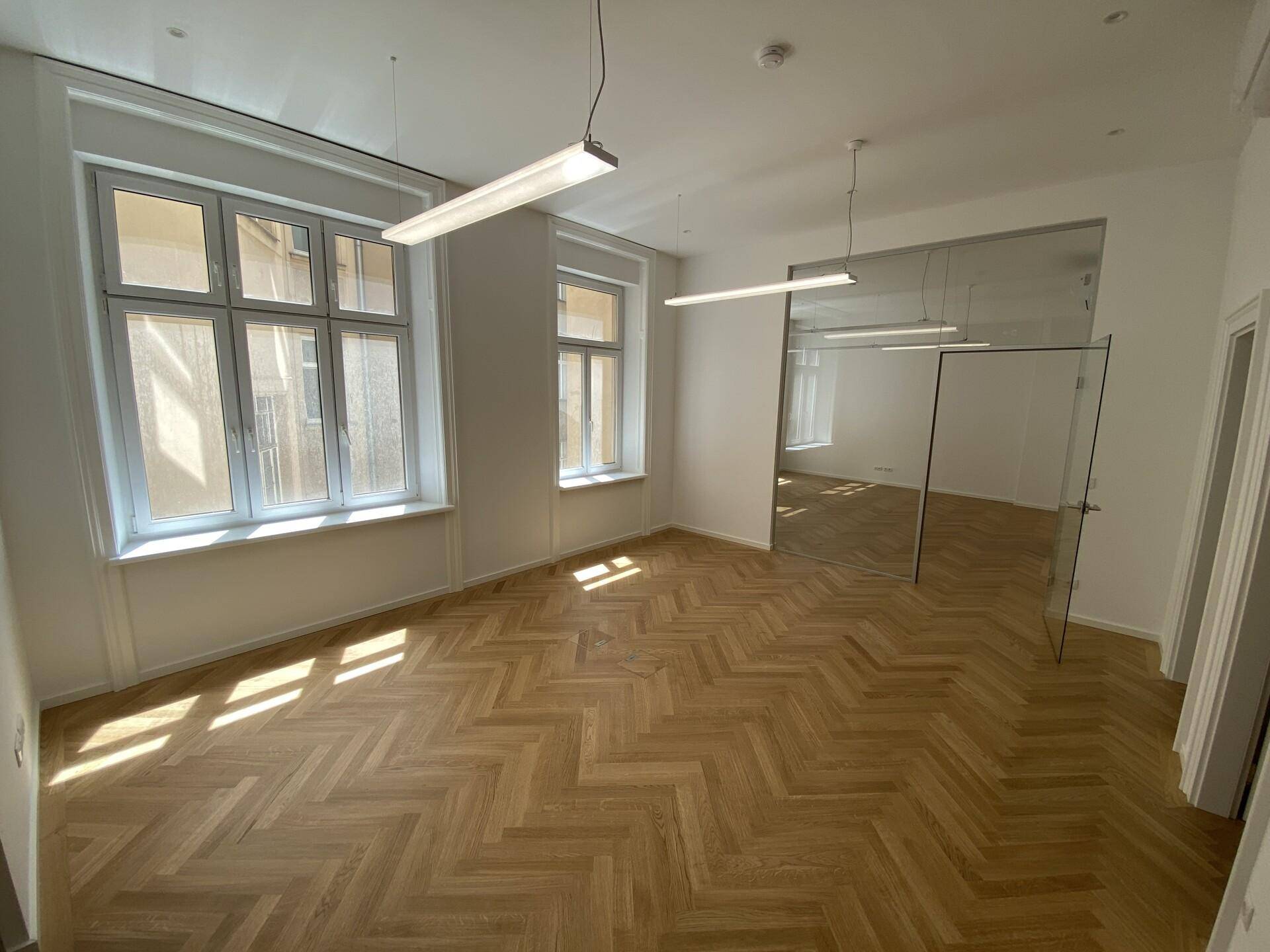 Helles Büro mit Fischgrätparkett, großen Fenstern und einer modernen Glaswand, die den Raum unterteilt.
