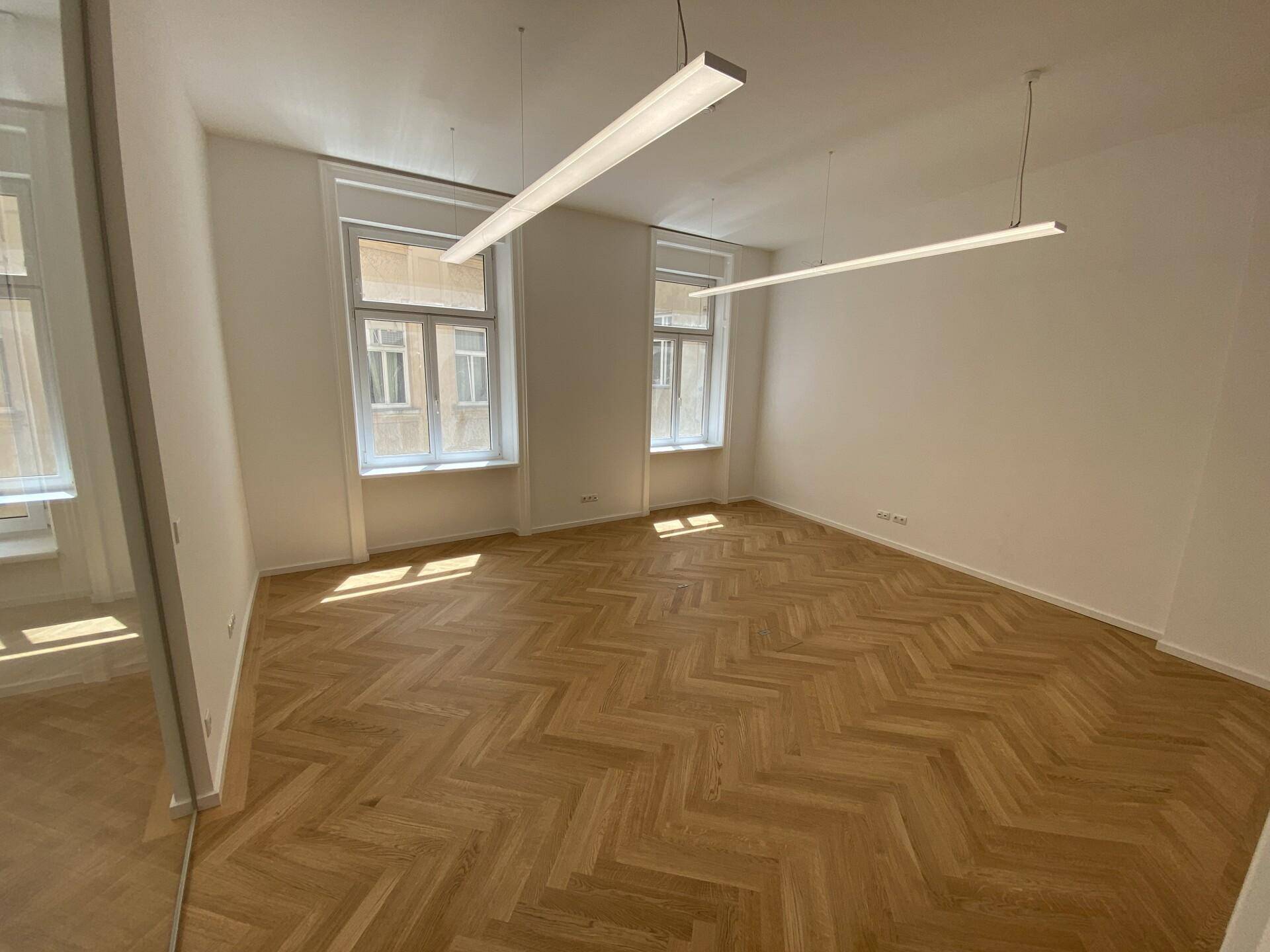 Heller Büroraum mit elegantem Fischgrätparkett und zwei großen Fenstern, die für viel natürliches Licht sorgen.