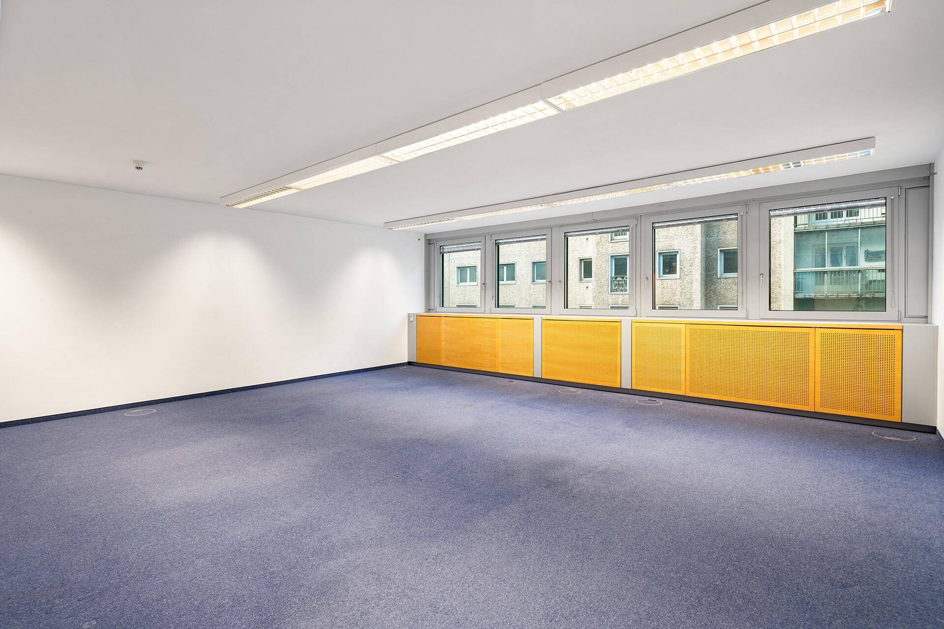 Leerer Büroraum mit blauem Teppichboden, weißen Wänden und gelben Heizungsverkleidungen unter den Fenstern.