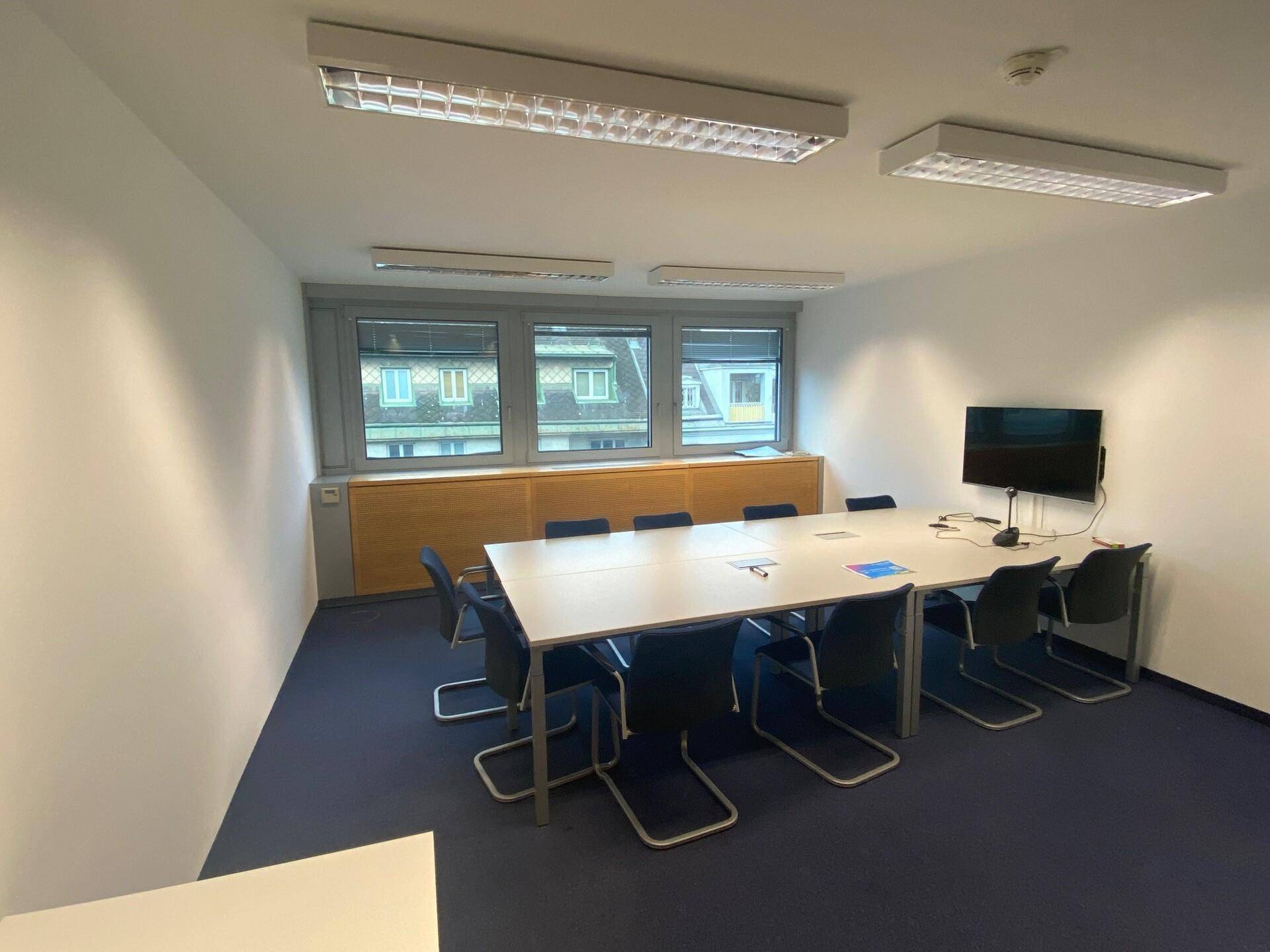 Konferenzraum mit rechteckigem Tisch, blauen Stühlen, kleinem Bildschirm und Blick auf die Stadt.