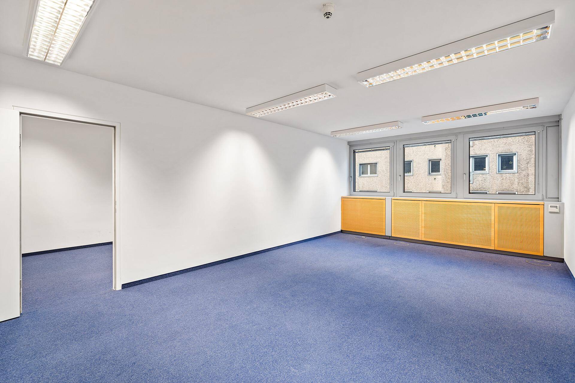 Leerer Büroraum mit blauem Teppichboden, weißen Wänden und gelben Heizungsverkleidungen unter den Fenstern.