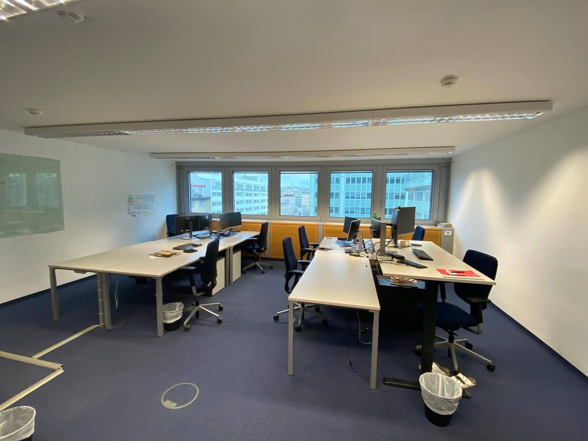 Geräumiger Großraumbüro mit mehreren Arbeitsplätzen, Computern und Fenstern, die Tageslicht hereinlassen.