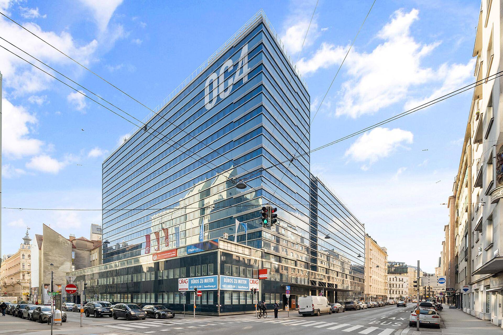 Modernes Bürogebäude mit beeindruckender Glasfassade an einer belebten Stadtstraße unter blauem Himmel.