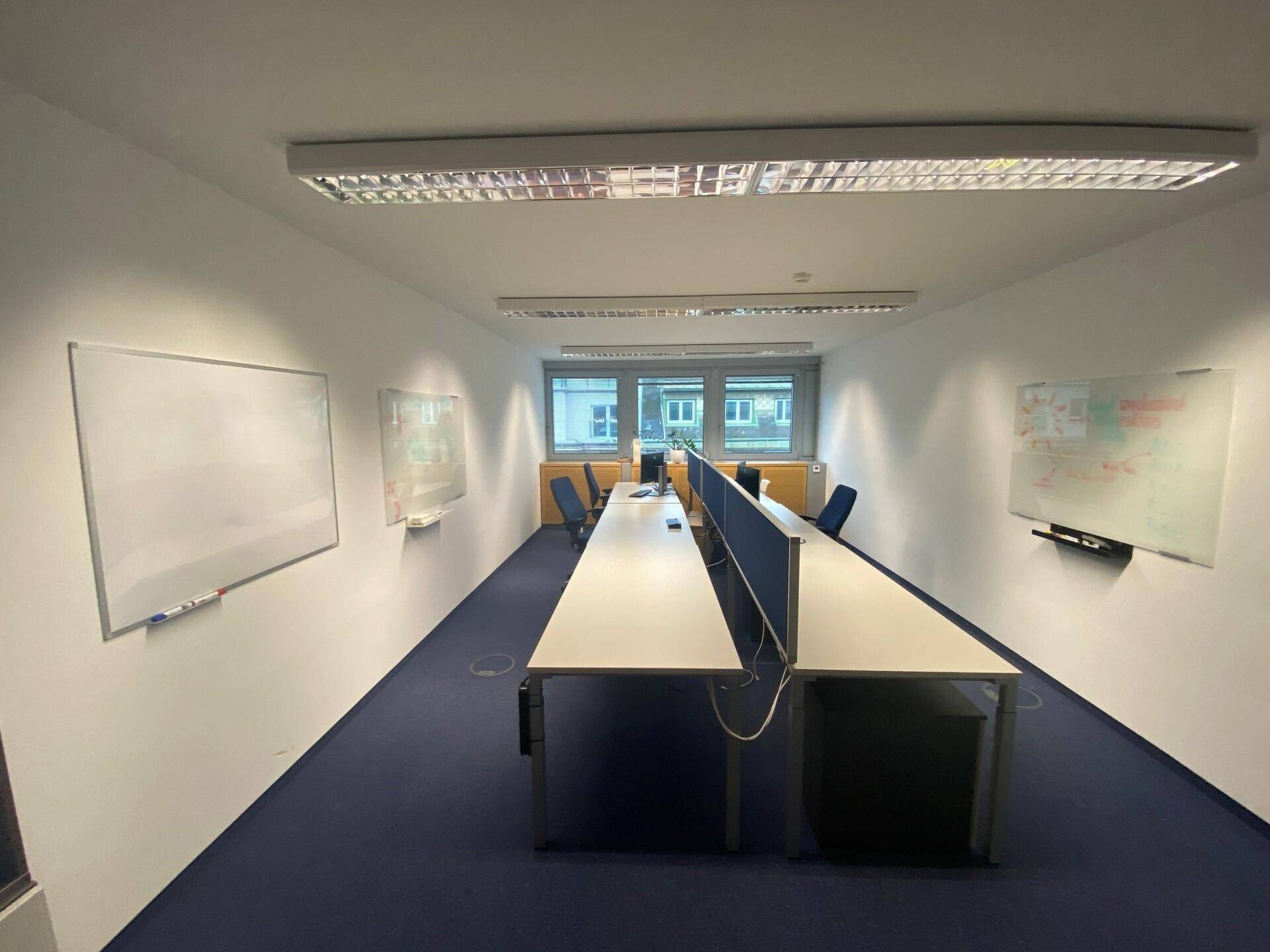Großraumbüro mit mehreren Arbeitsplätzen, Whiteboards und Fenstern, die eine kollaborative Umgebung fördern.