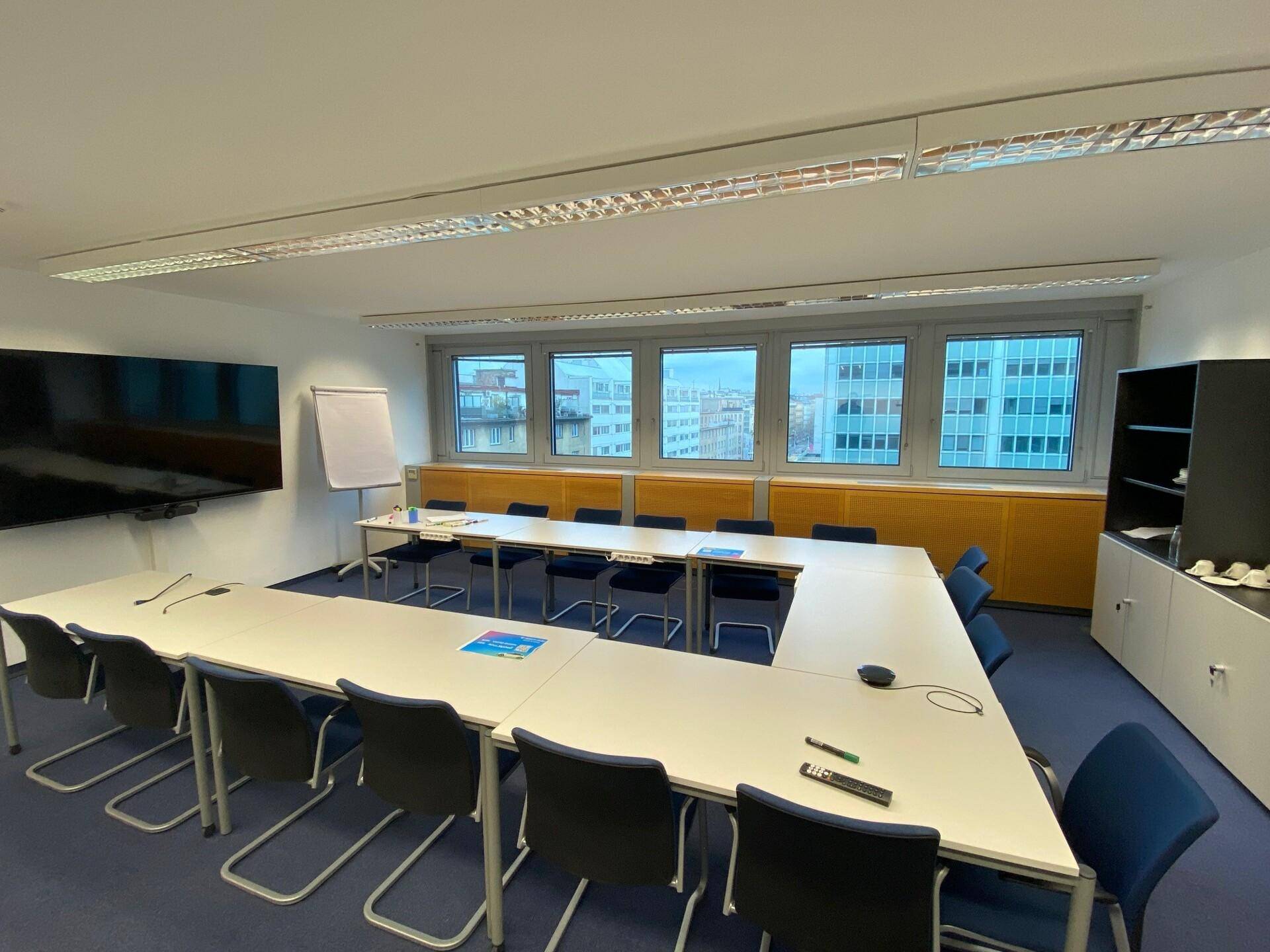 Großer Konferenzraum mit U-förmigem Tisch, blauer Teppichboden und Fenstern mit Stadtblick.