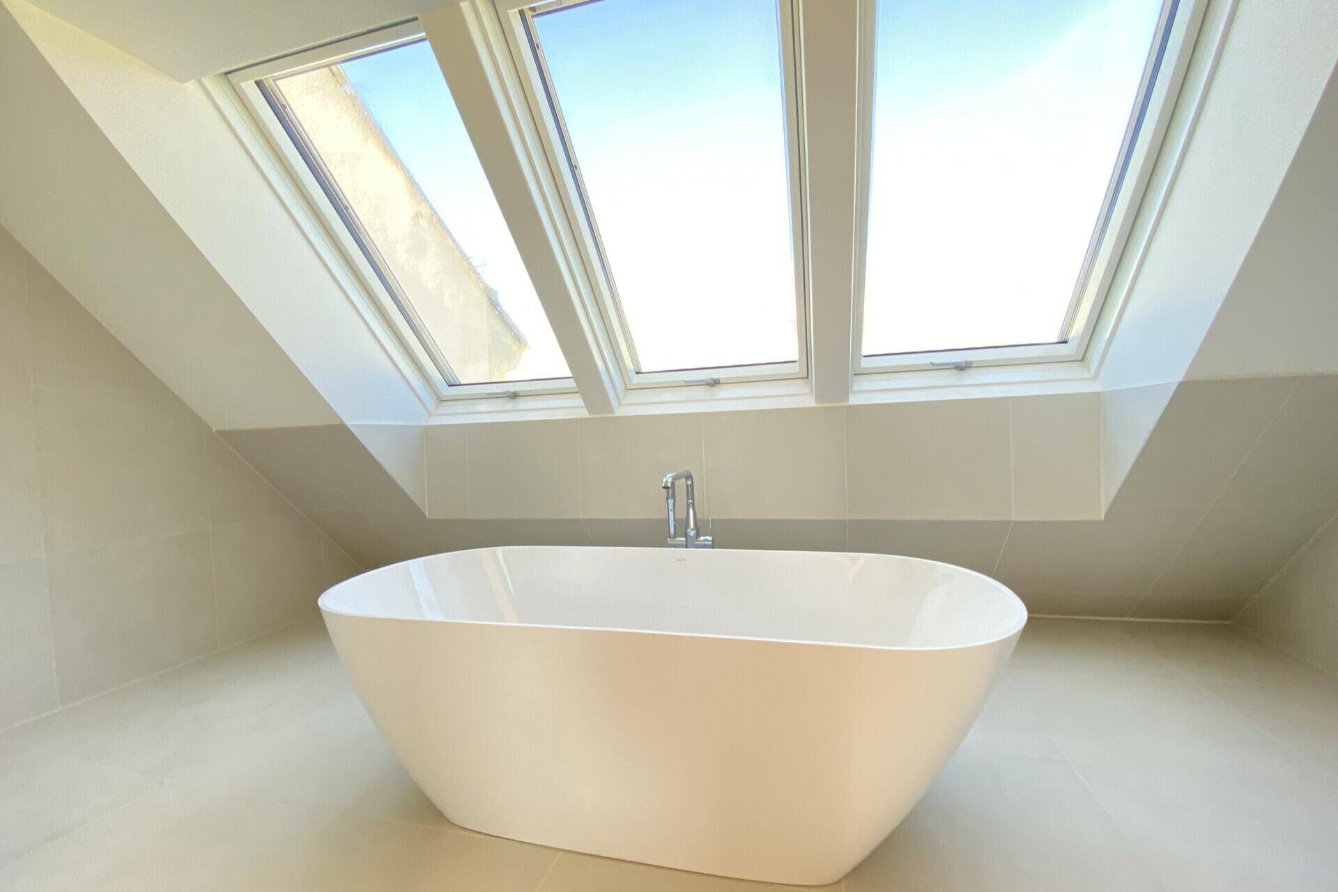 Modernes Badezimmer mit freistehender Badewanne unter drei Dachfenstern und hellen Fliesen.