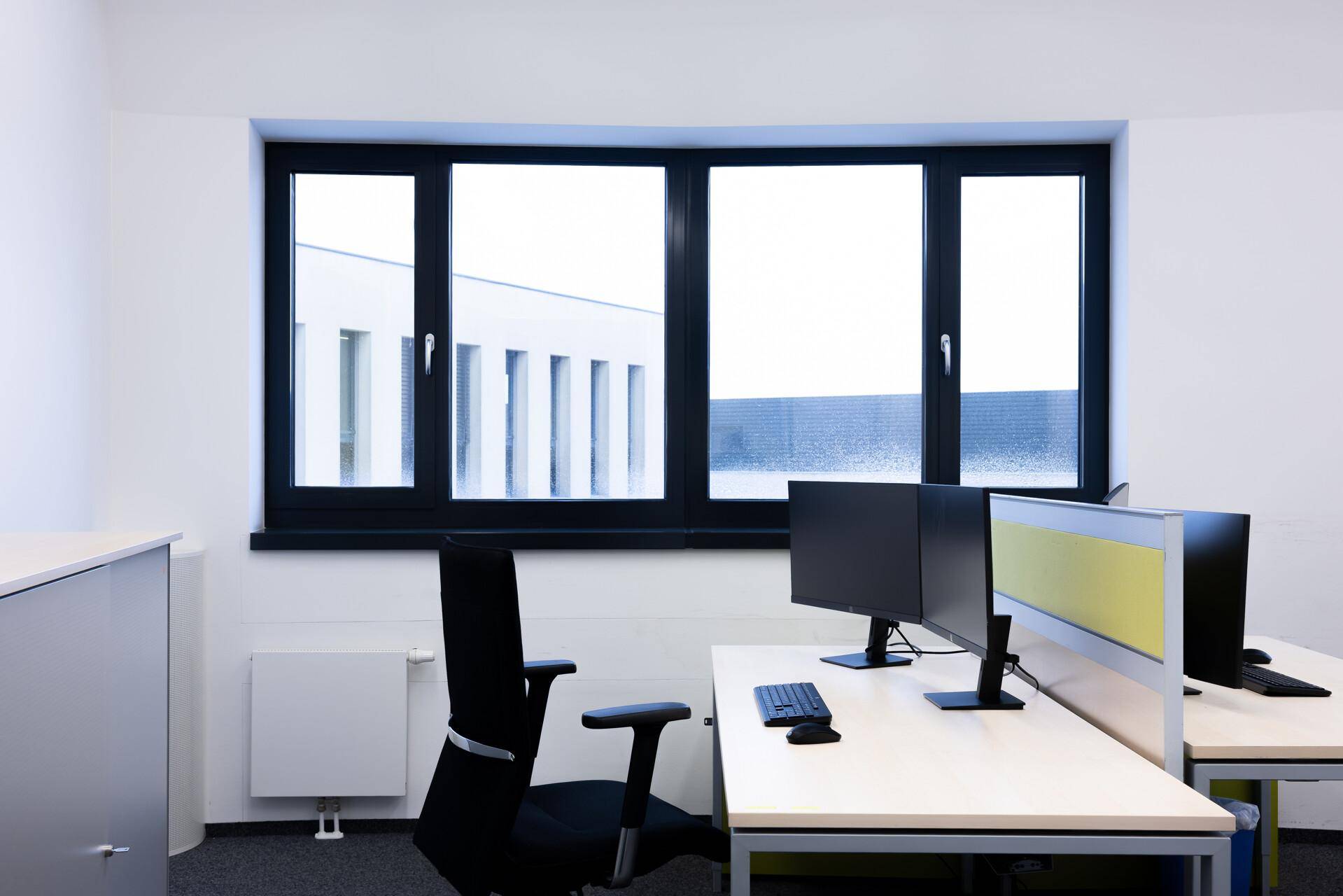 Büroraum mit zwei Arbeitsplätzen, Stühlen und Computermonitoren vor einem großen Fenster.