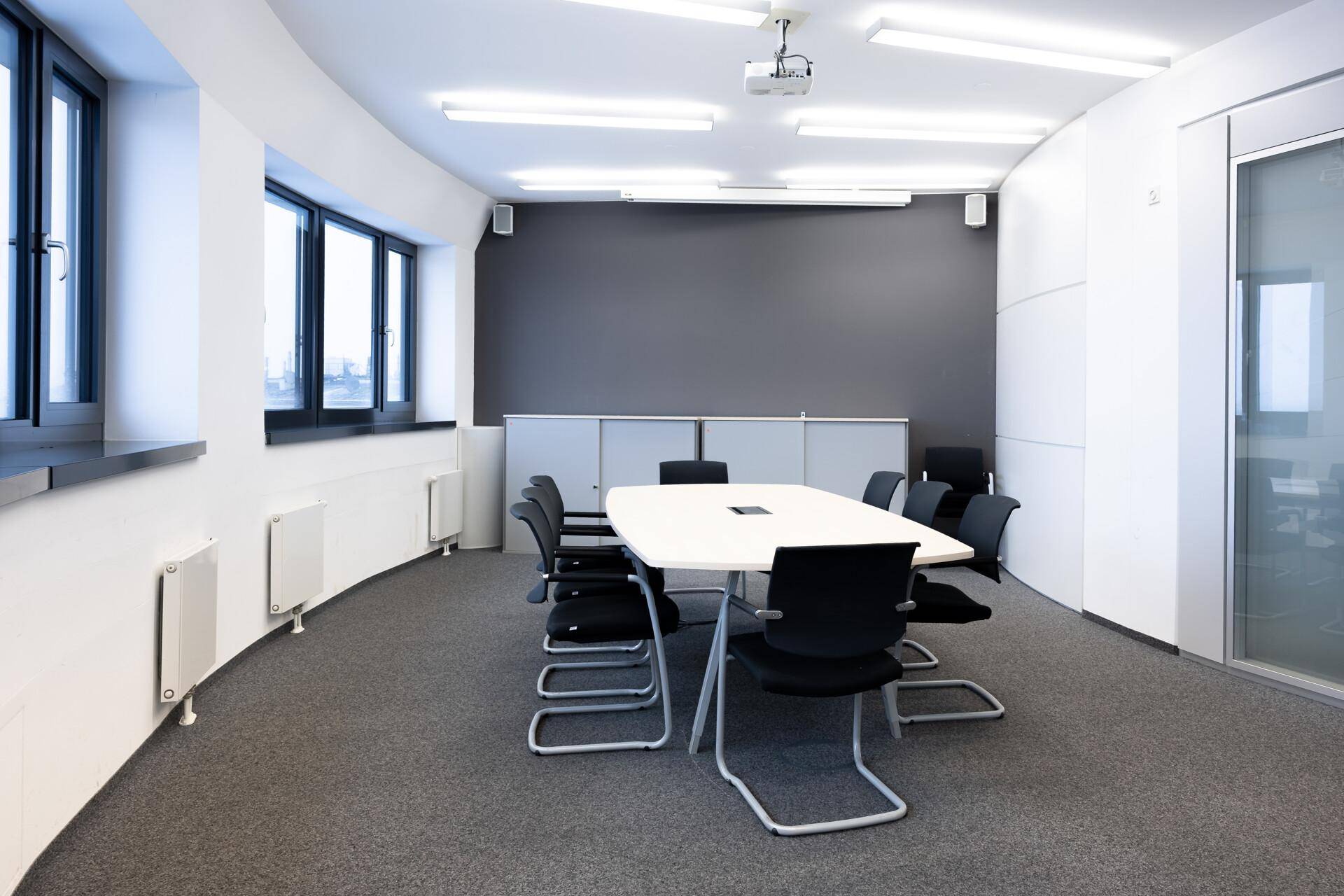 Moderner Konferenzraum mit großem weißen Tisch, schwarzen Stühlen, Beamer und einer geschwungenen Fensterwand.