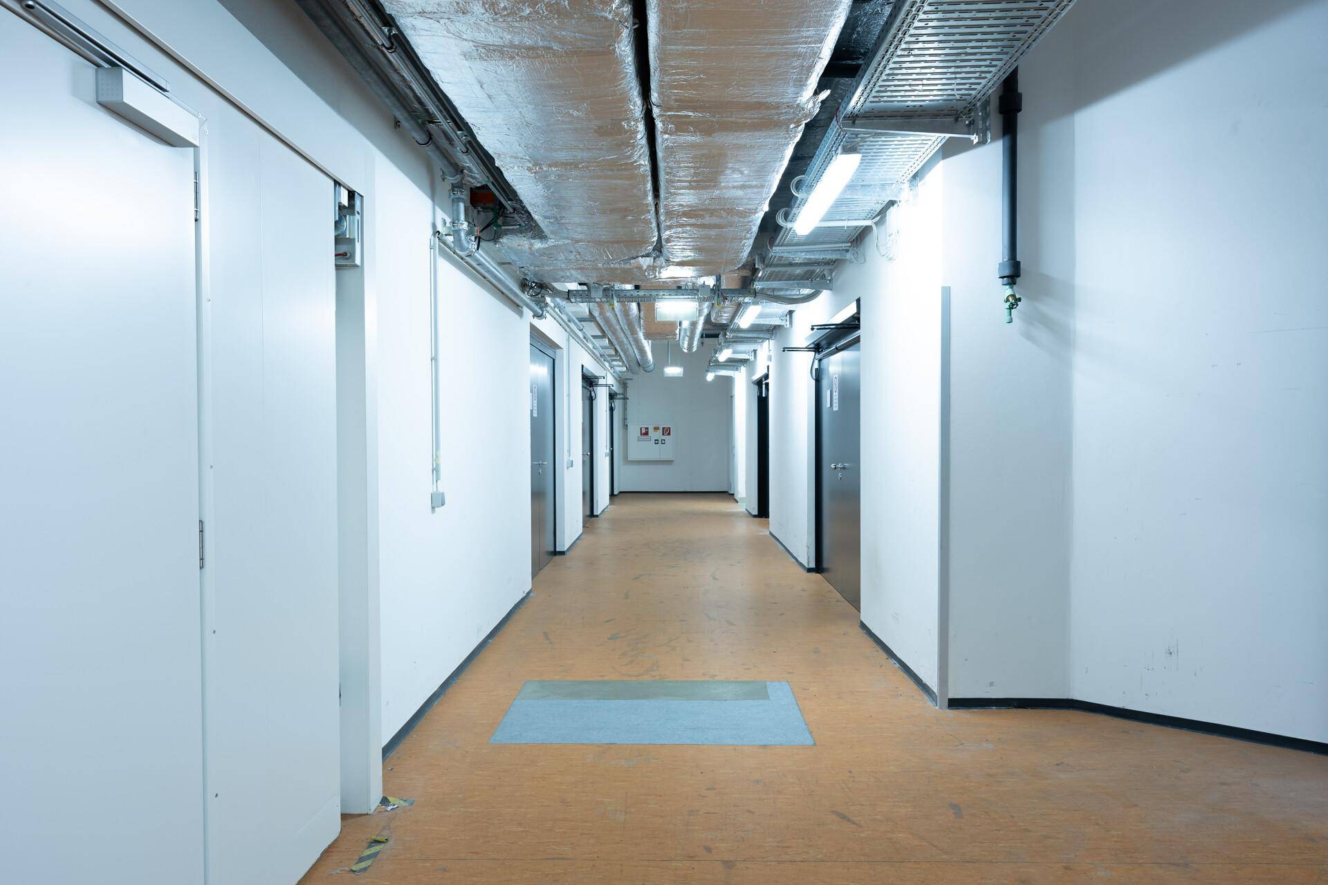 Langer, leerer Flur mit weißen Wänden, hellem Boden und sichtbaren Rohren und Kanälen an der Decke.
