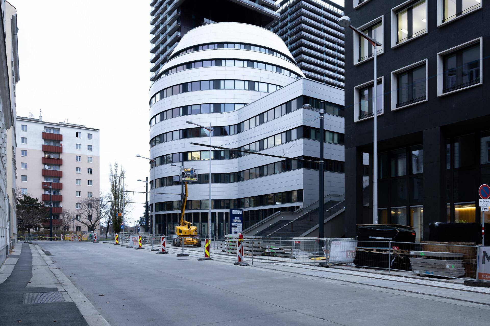 Straßenansicht mit einem modernen, geschwungenen Gebäude, umgeben von anderen Bauten und Baustellenelementen.