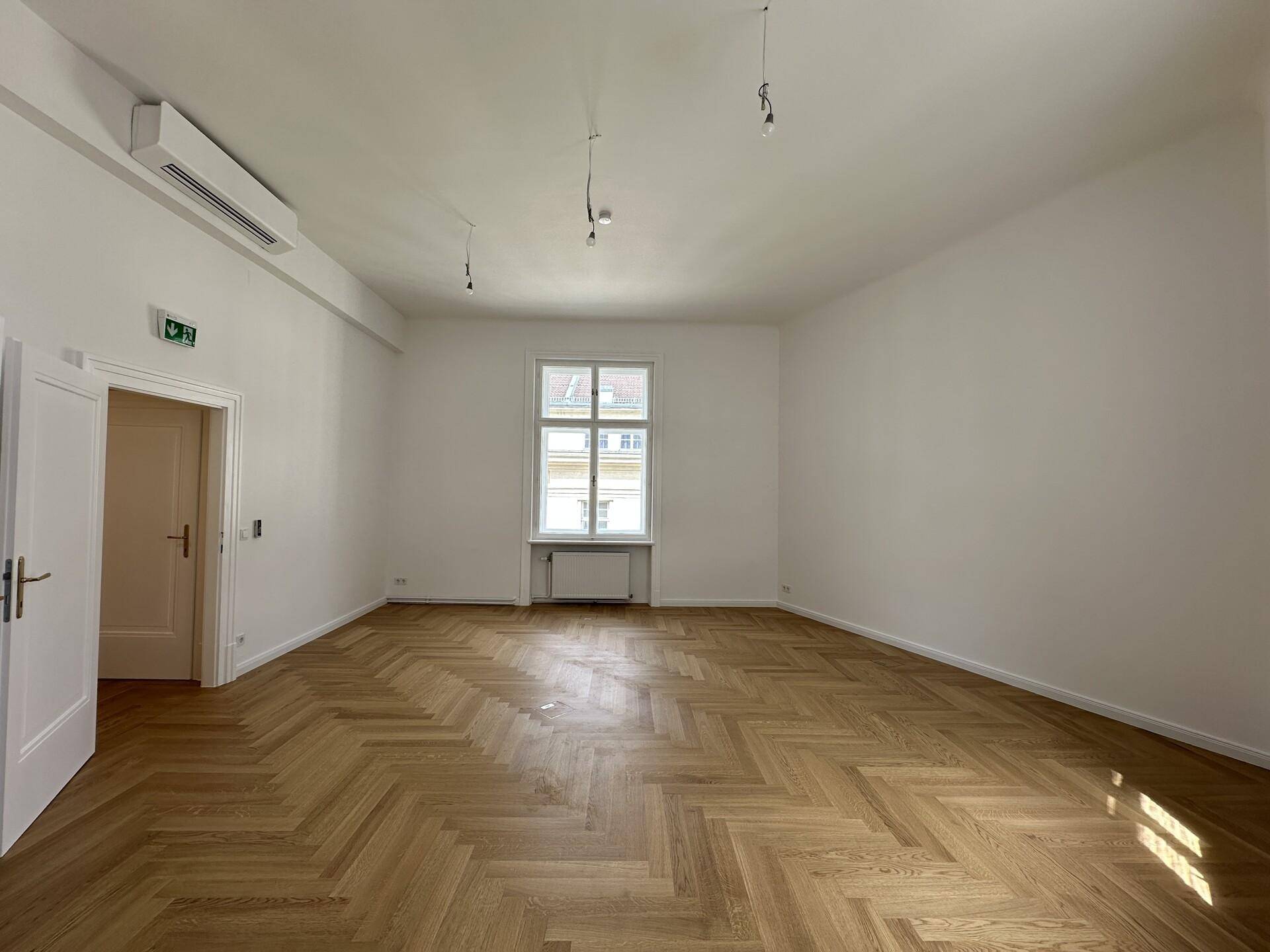 Heller Raum mit Fischgrätparkett, einem großen Fenster und Klimaanlage an der Wand.