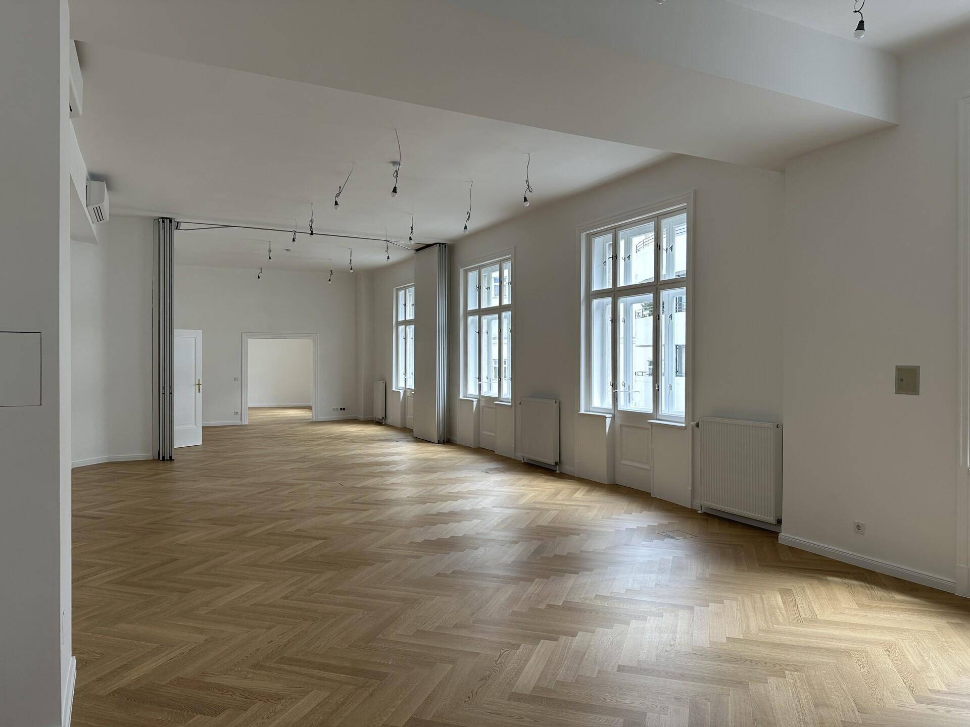 Geräumiger, heller Raum mit Fischgrätparkett, hohen Fenstern und moderner Beleuchtung.