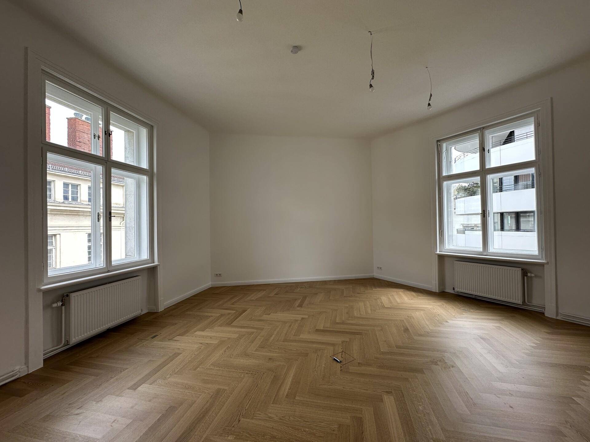 Großer, heller Raum mit Fischgrätparkett und zwei hohen Fenstern mit Blick nach draußen.