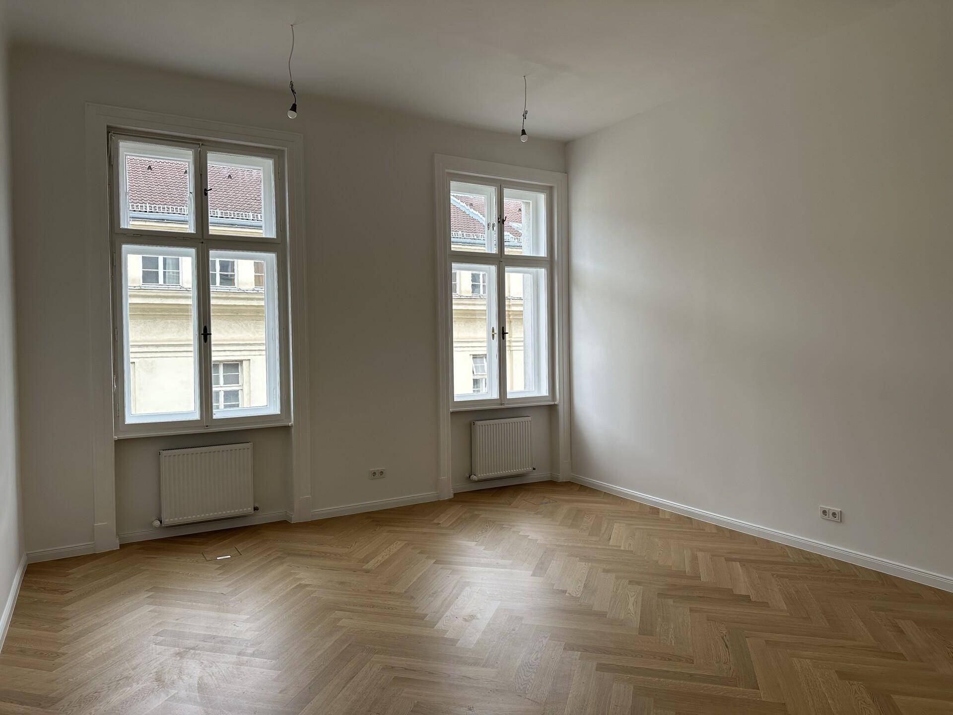 Heller Raum mit Fischgrätparkett und zwei hohen Fenstern, die viel Tageslicht hereinlassen.