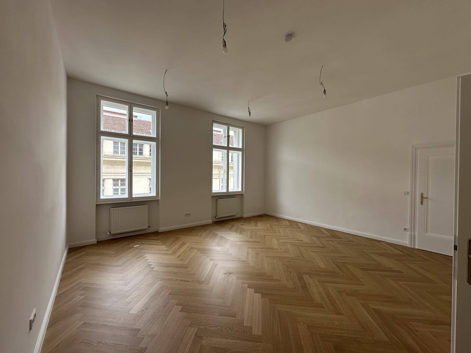 Heller Raum mit Fischgrätparkett und zwei hohen Fenstern, ideal für Wohn- oder Arbeitszwecke.