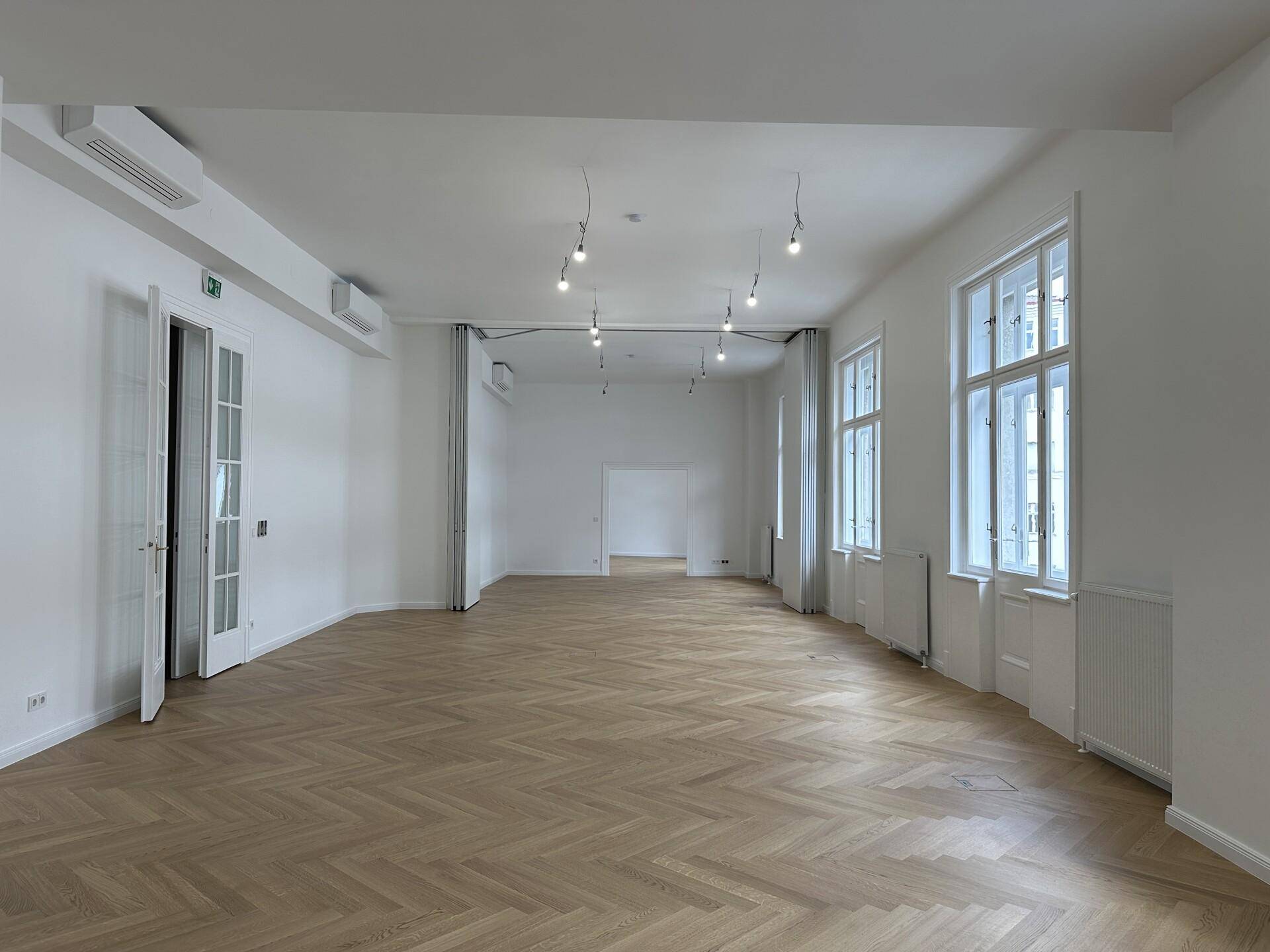 Großer, heller Raum mit Fischgrätparkett, hohen Fenstern und Klimaanlage.