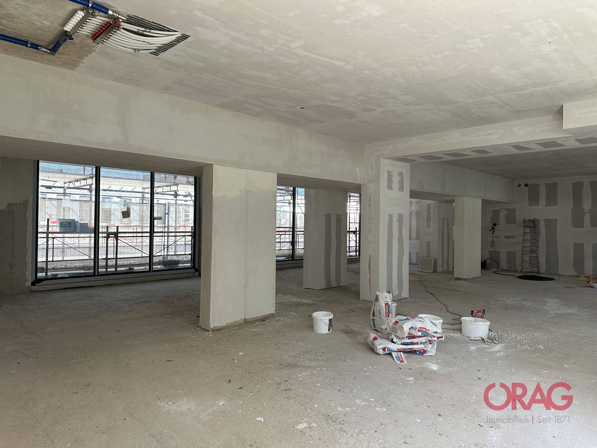 Weitläufiger Innenbereich im Rohbau mit hohen Decken und Baumaterialien auf dem Betonboden.