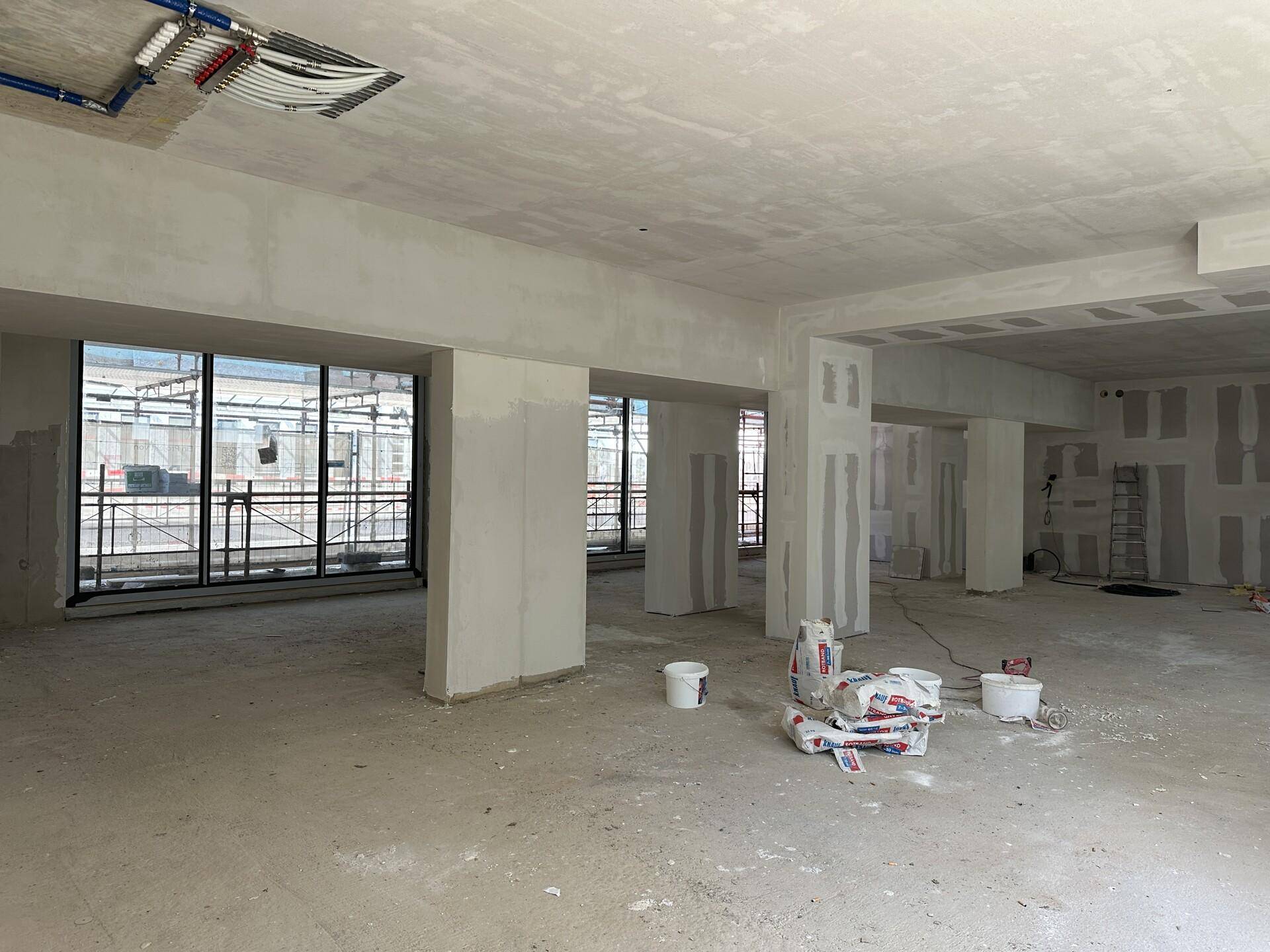 Geräumiger, unfertiger Innenraum mit hohen Decken und Blick auf die Baustelle durch große Fenster.