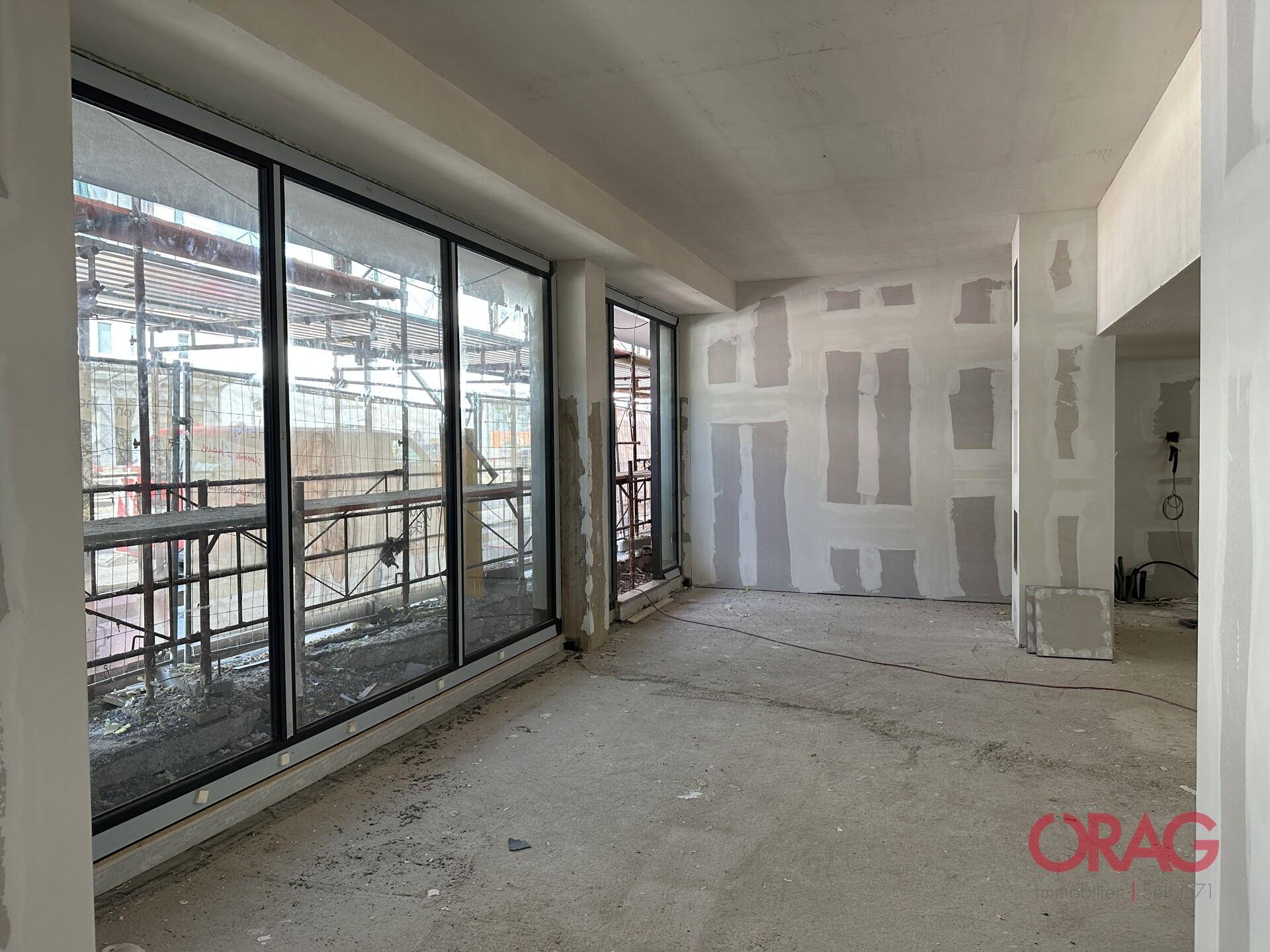 Weitläufiger Innenraum im Rohbau mit großen Fensterfronten und Blick auf das Baugerüst.