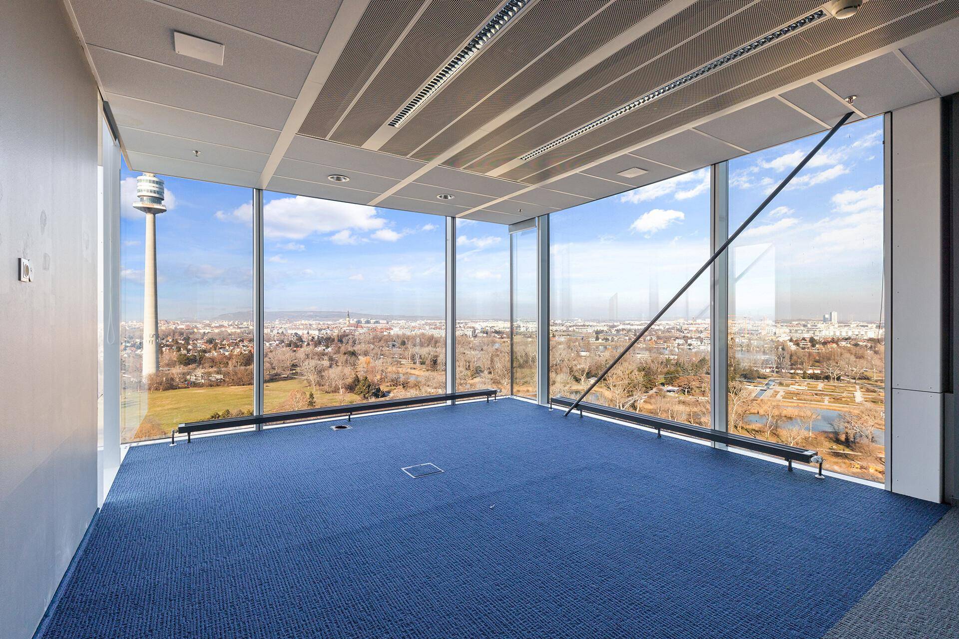 Leerer Büroraum mit großen Fenstern, blauem Teppichboden und weitem Blick über die Stadt.