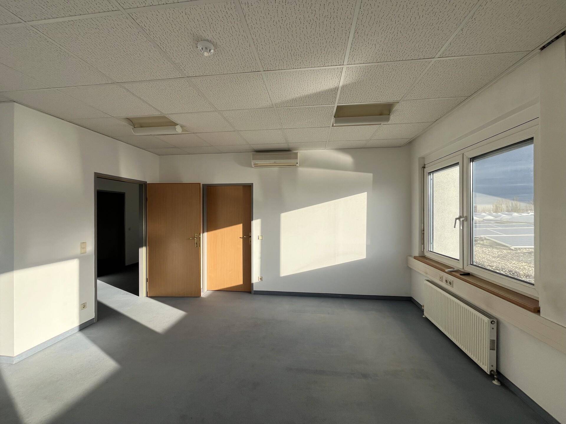 Leerer Büroraum mit weißen Wänden, dunklem Boden, Rasterdecke und zwei Holztüren, Klimaanlage sichtbar.