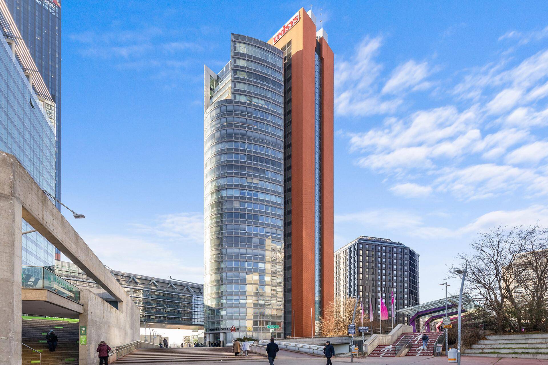 Modernes Hochhaus mit geschwungener Glasfassade und angrenzendem Turm unter blauem Himmel.