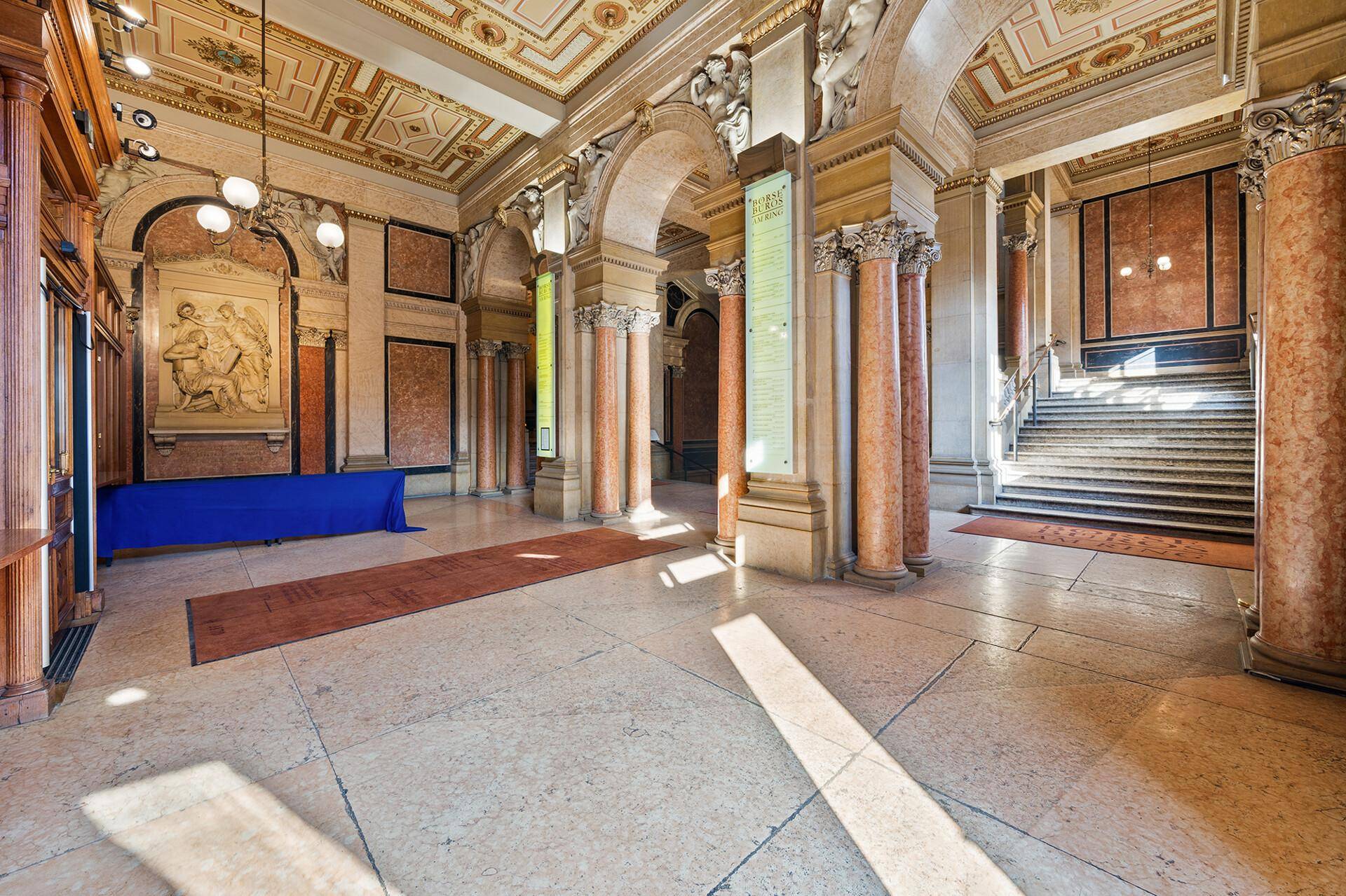 Historische Halle mit verzierten Decken, Marmorsäulen und einer breiten Treppe im Hintergrund.